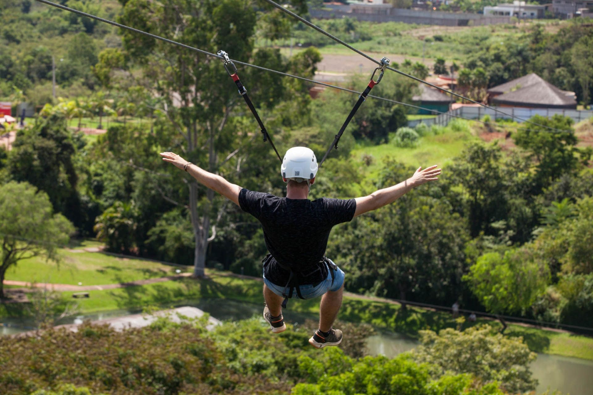 Pessoa deslizando em tirolesa sobre um vale verde com os braços estendidos e usando capacete.