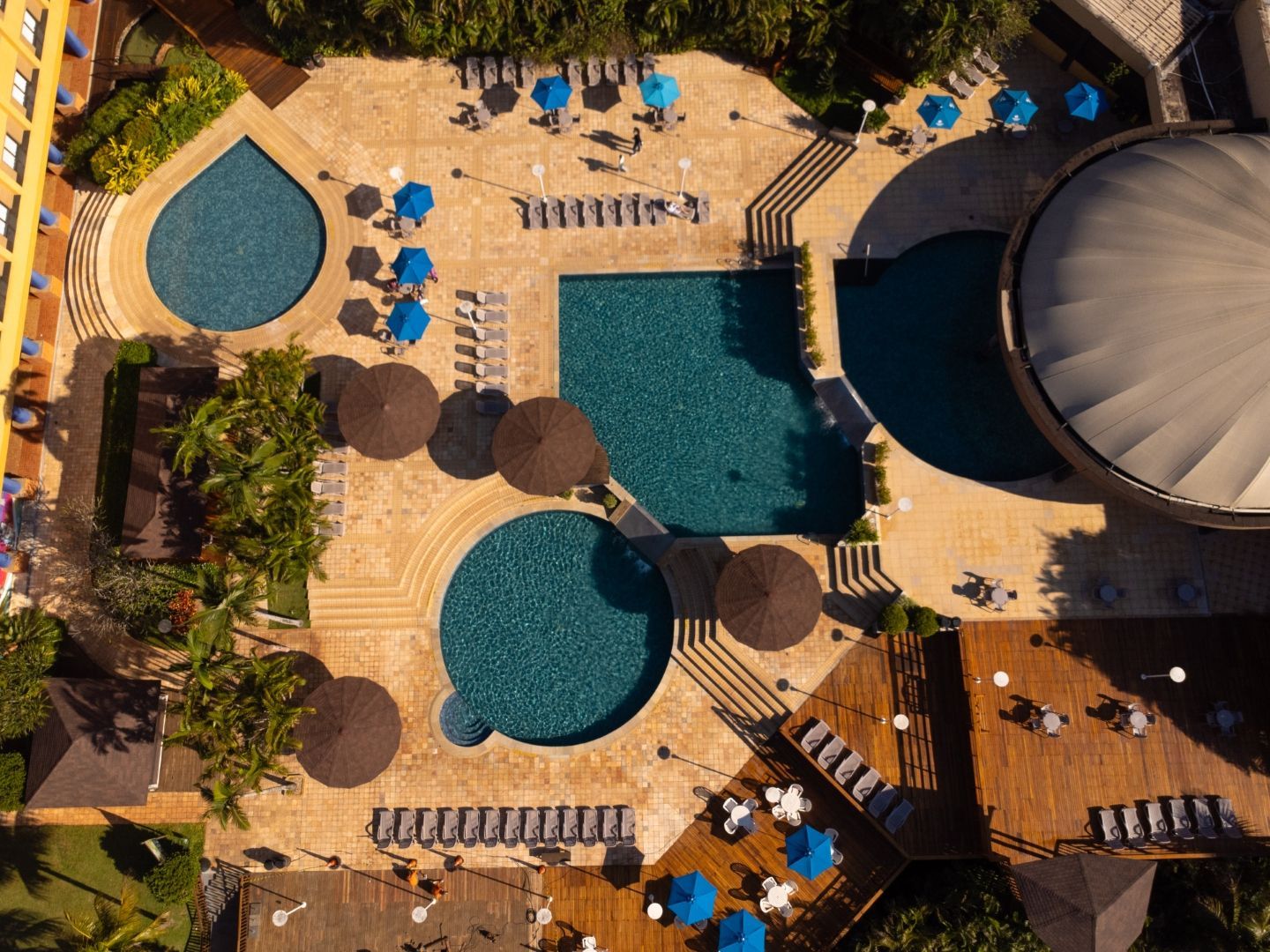 Vista aérea da área da piscina de um resort com piscinas de água turquesa, espreguiçadeiras, guarda-sóis e decks de madeira.