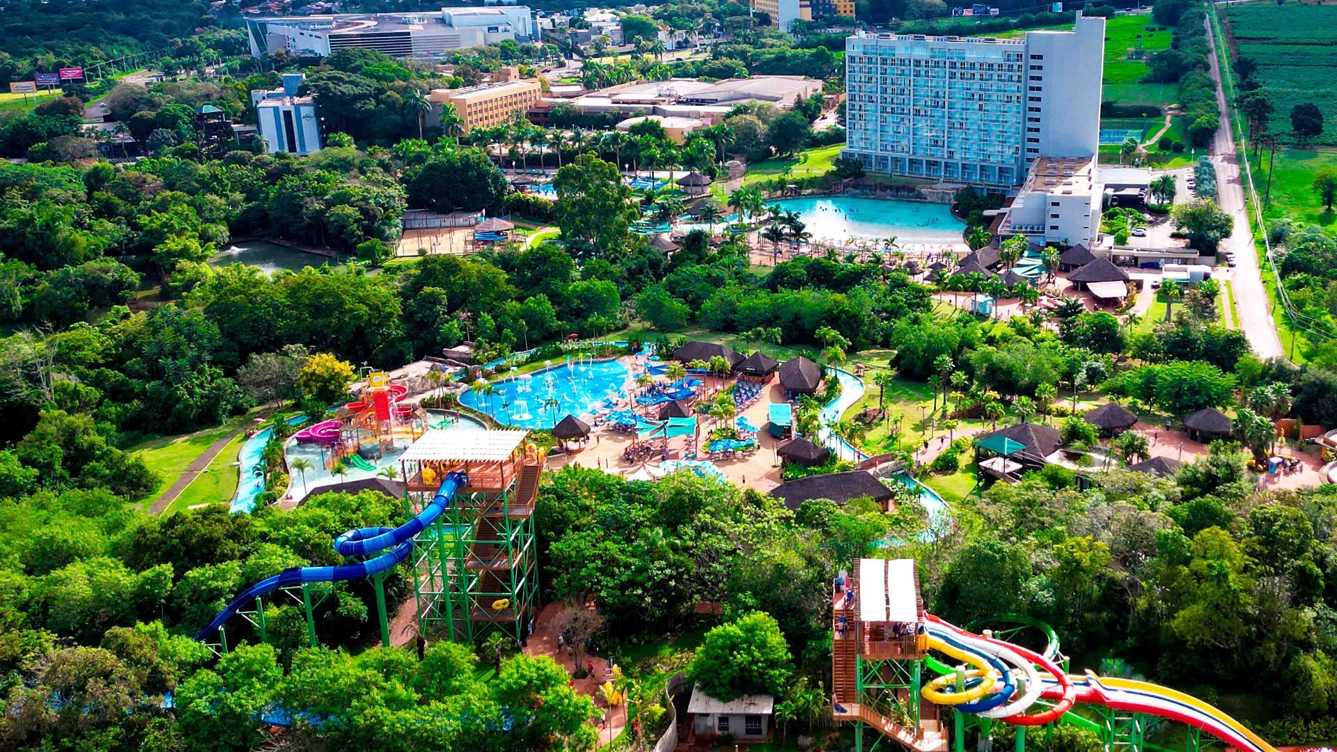 Vista aérea de um resort exuberante com piscinas, toboáguas e um hotel alto cercado por vegetação densa.