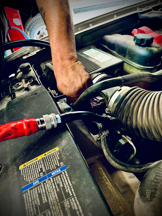 Person's hand working on a car engine. Black hose with red covering attached to the engine bay.