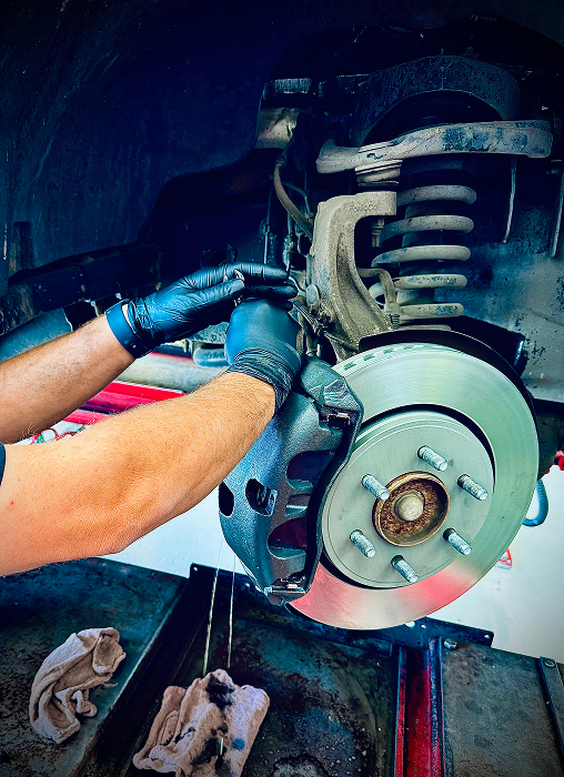 Mechanic replacing brake caliper on a car wheel; gloved hands, rotor, suspension. | King's Auto LLC