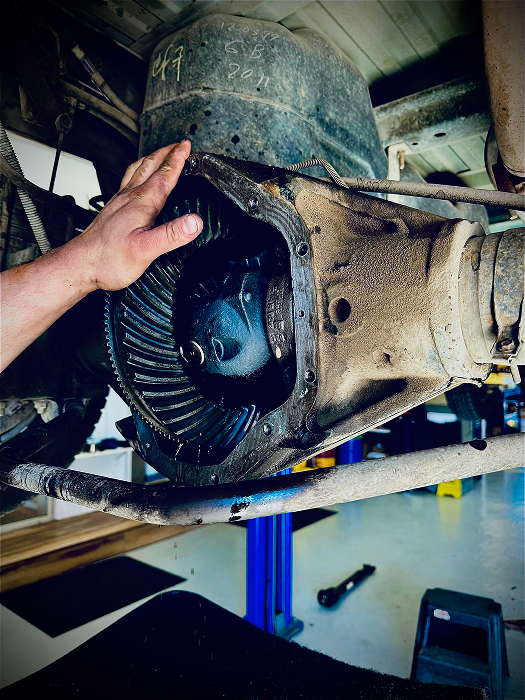 Hand pointing at an open differential housing on a vehicle lift, revealing gears and oil. | King's Auto LLC