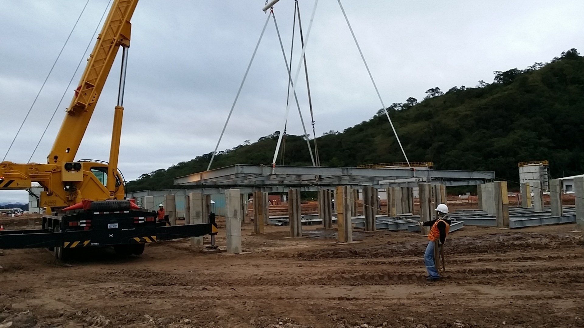 A yellow crane lifts a large metal beam onto concrete pillars at a construction site against a hillside.