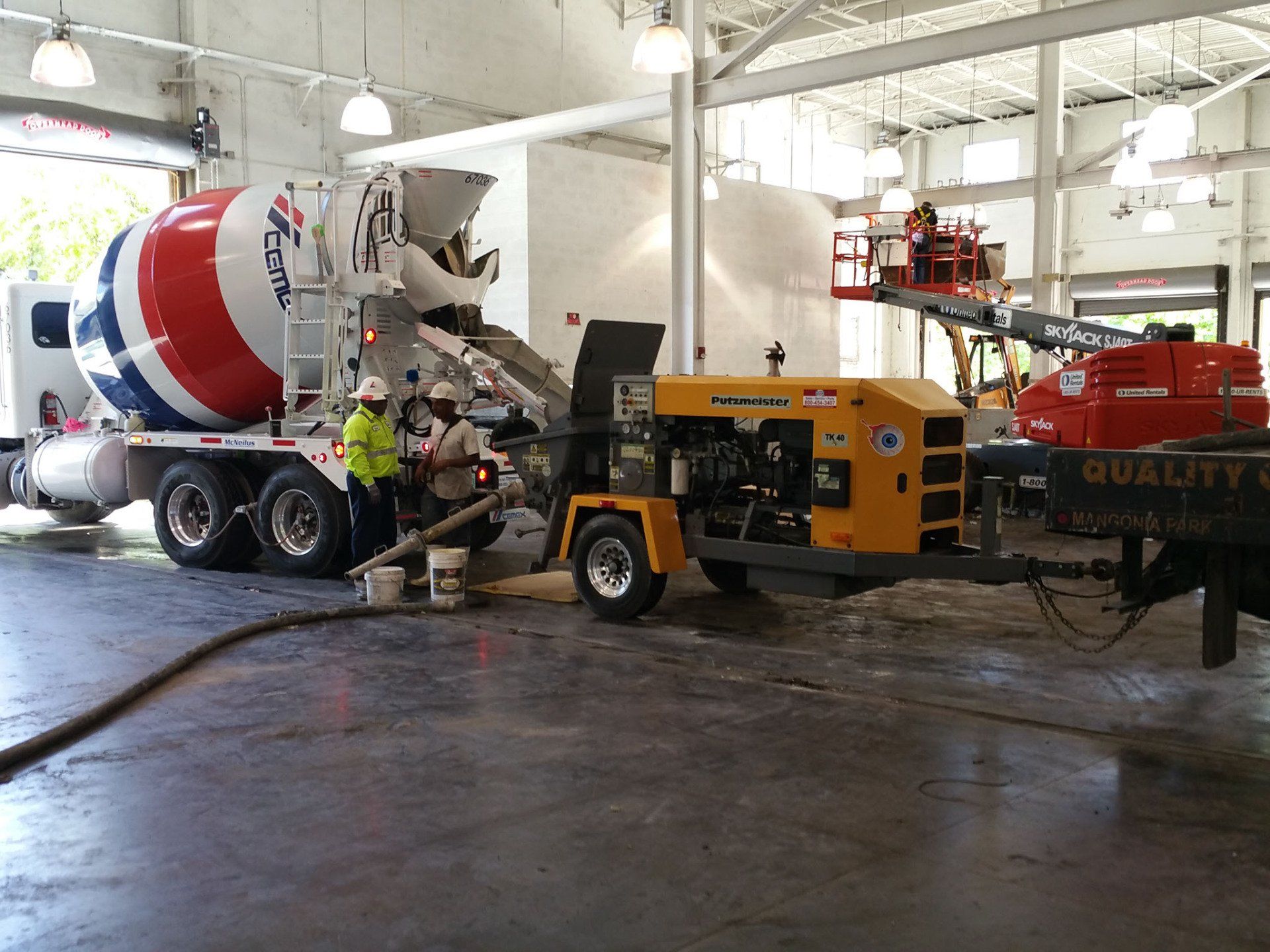 Workers pour wet concrete from a red, white, and blue truck into a yellow industrial pump inside a warehouse.