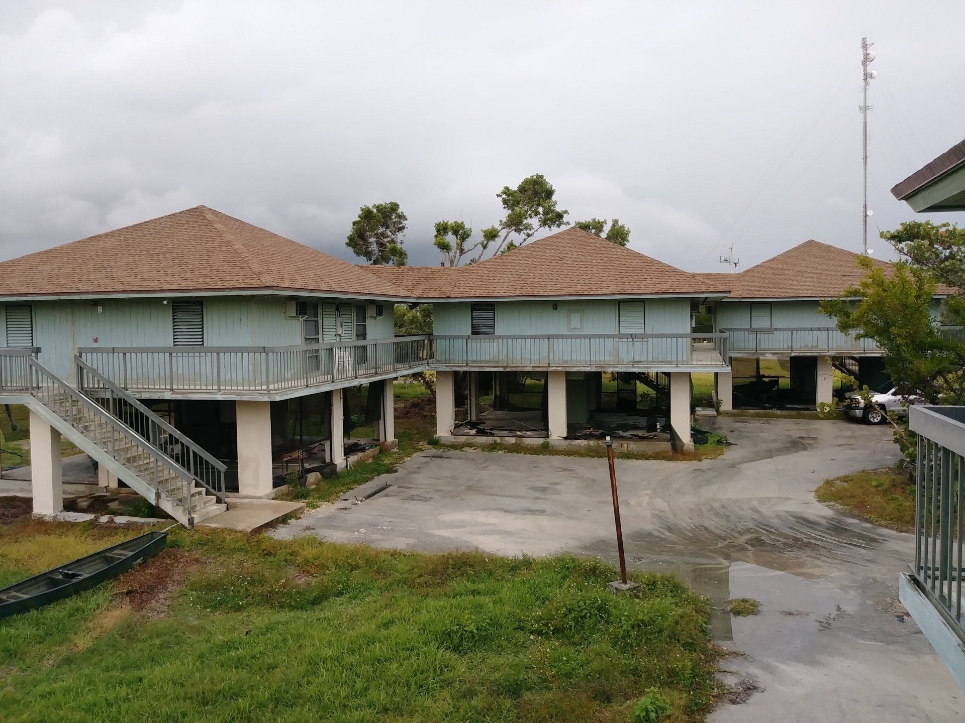 Three elevated, light-blue stilt houses with brown shingled roofs sit on a paved area under a cloudy sky.
