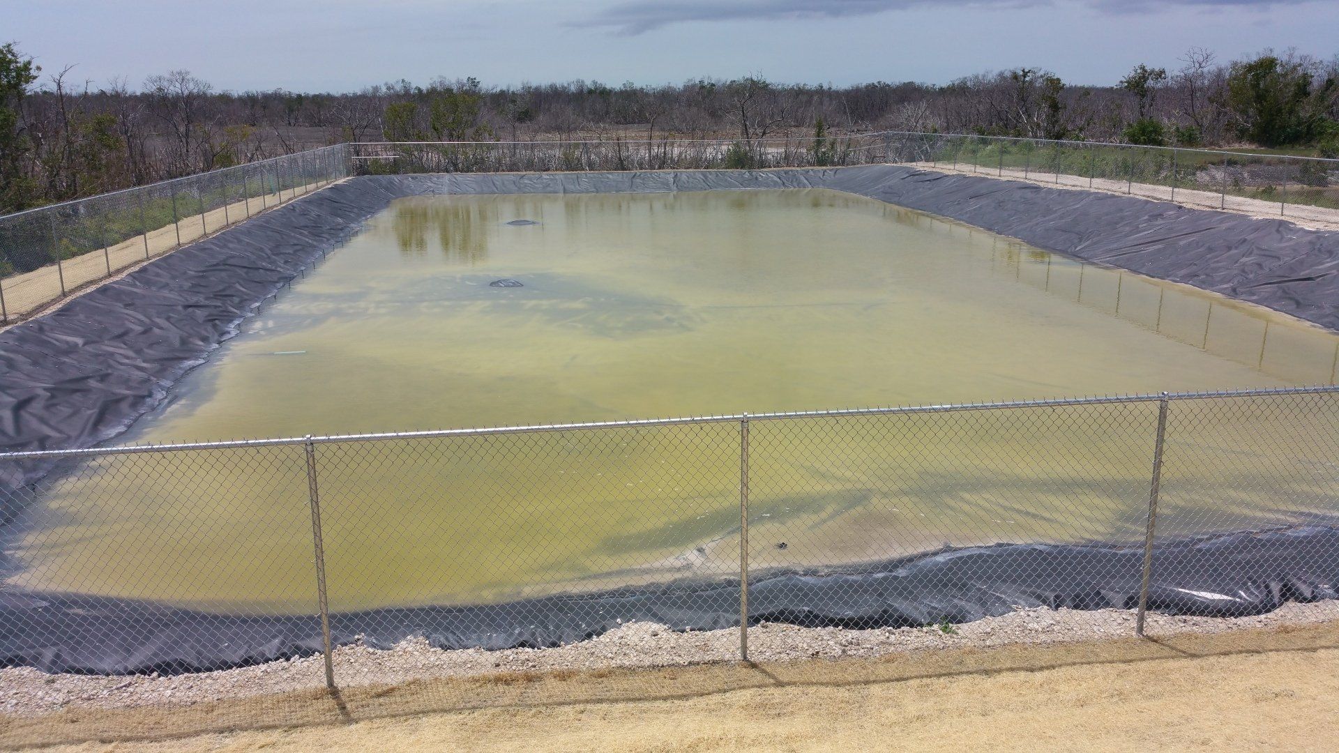 A large, rectangular water reservoir with a dark liner, surrounded by a chain-link fence in an open outdoor setting.