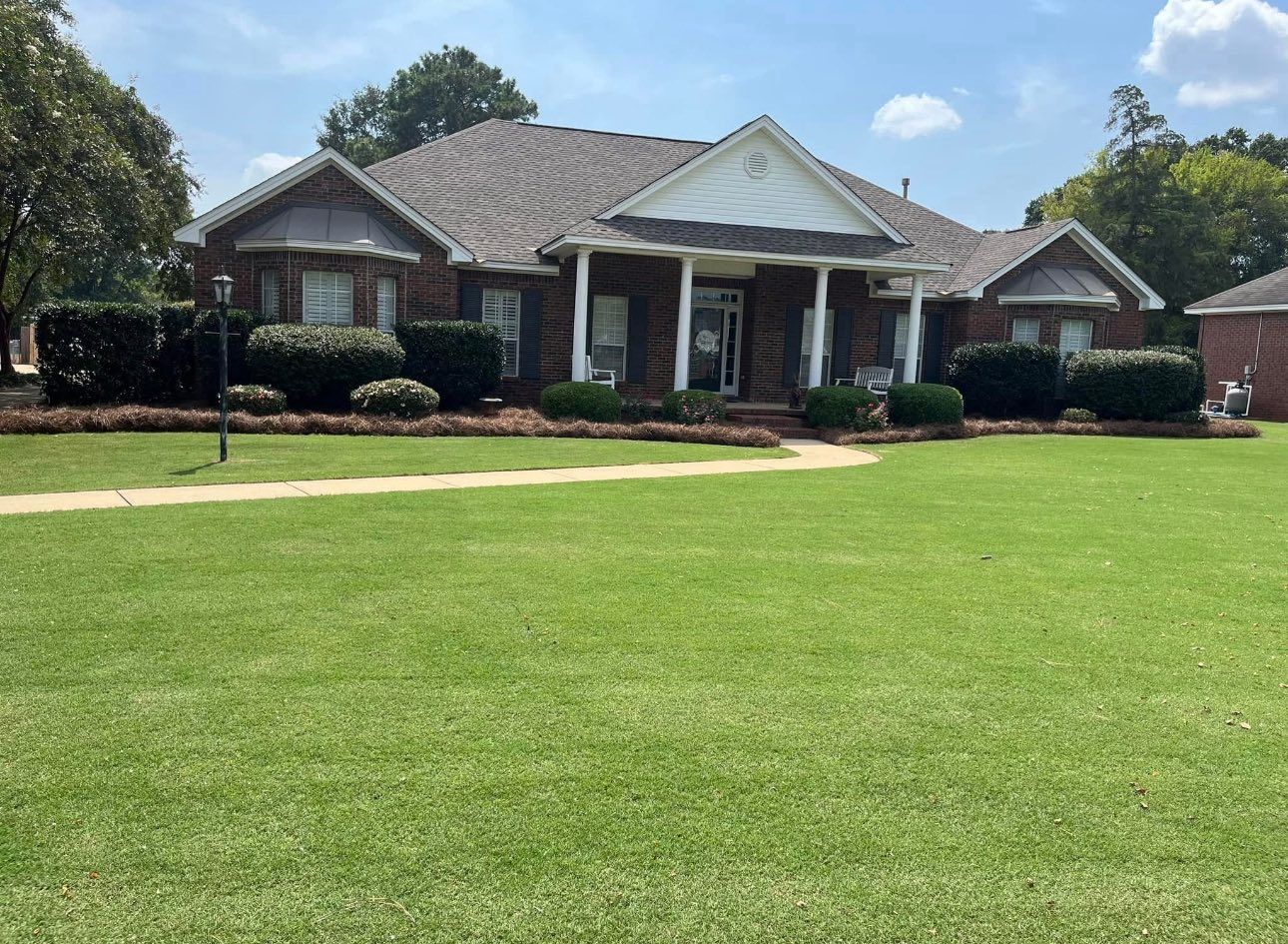 A one-story brick house with a white gabled porch and a sprawling green lawn under a bright blue sky.