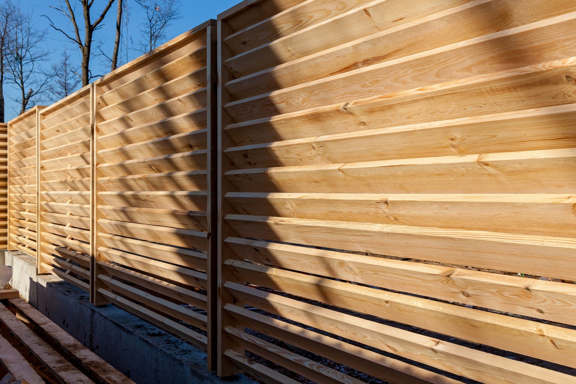 A modern wooden louvered fence made of horizontal cedar planks, mounted on a concrete base against a blue sky.
