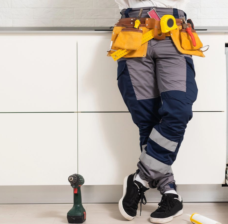 Una persona in pantaloni da lavoro è in piedi in cucina con una cintura portautensili, un trapano sul pavimento e un metro a nastro giallo.