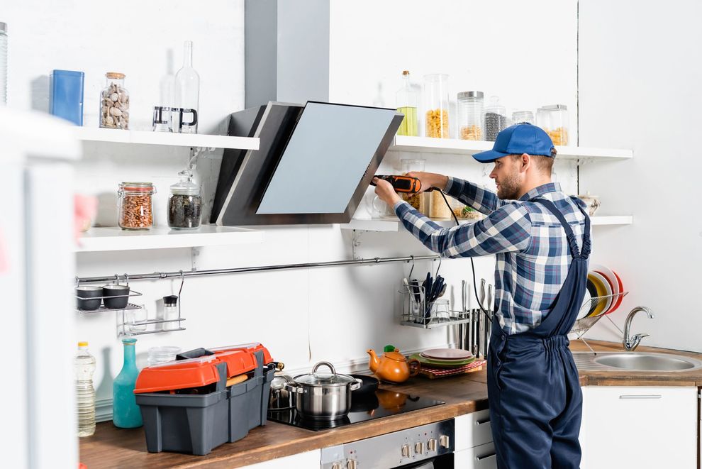 Un tecnico ripara un mobile da cucina sotto una cappa aspirante rialzata in una cucina moderna e luminosa.