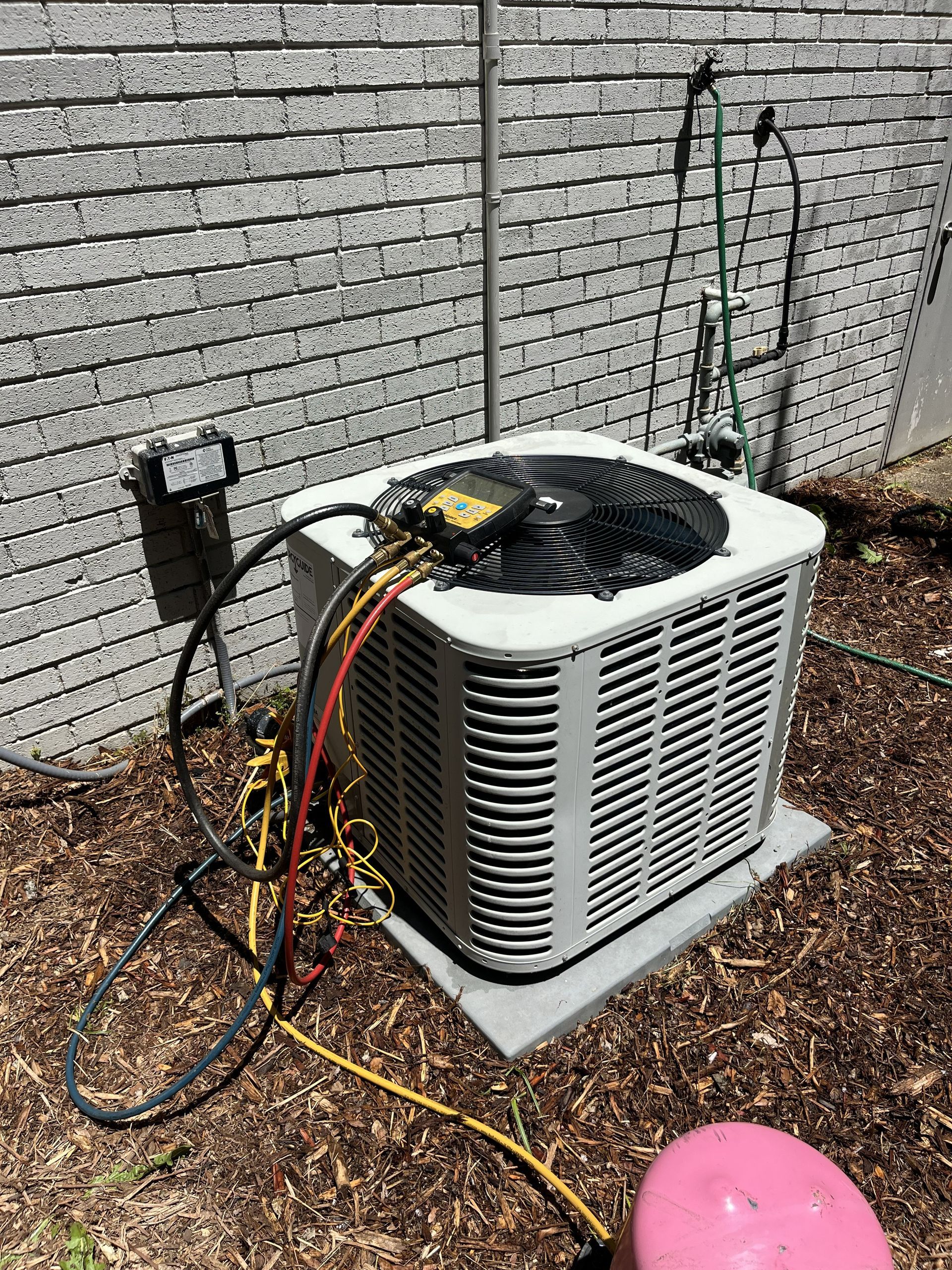 Outdoor HVAC condenser unit with service gauges and hoses attached beside a brick wall