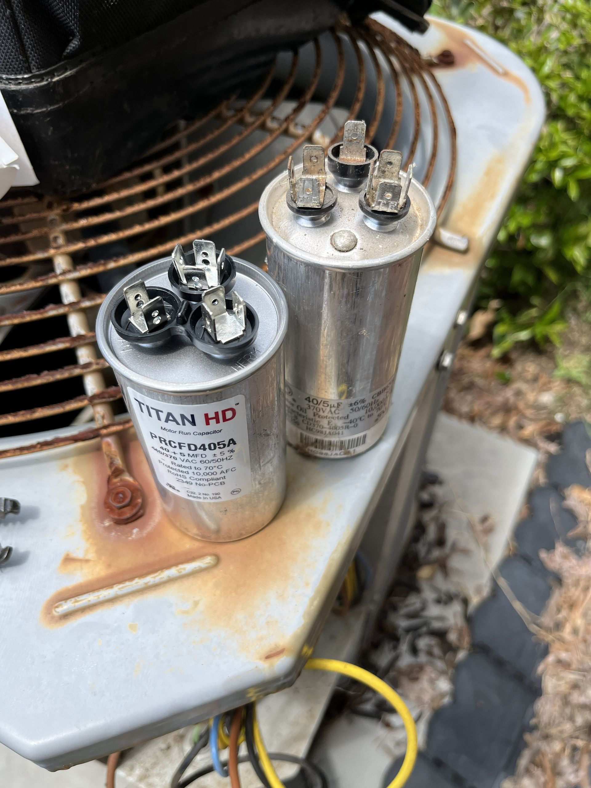 Two cylindrical HVAC capacitors on a rusted outdoor unit, with corroded wiring and a yellow cable nearby