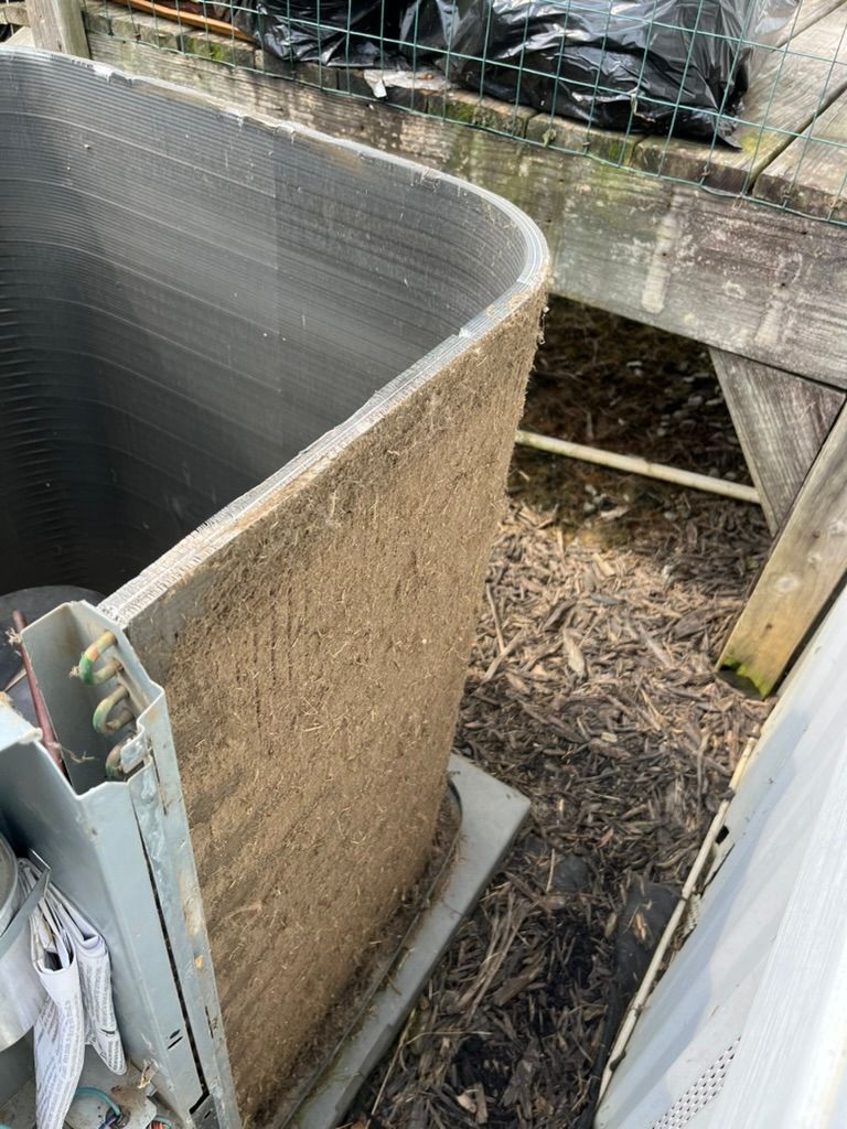 Outdoor HVAC condenser with a curved metal coil and tan insulation beside a wall and concrete support.