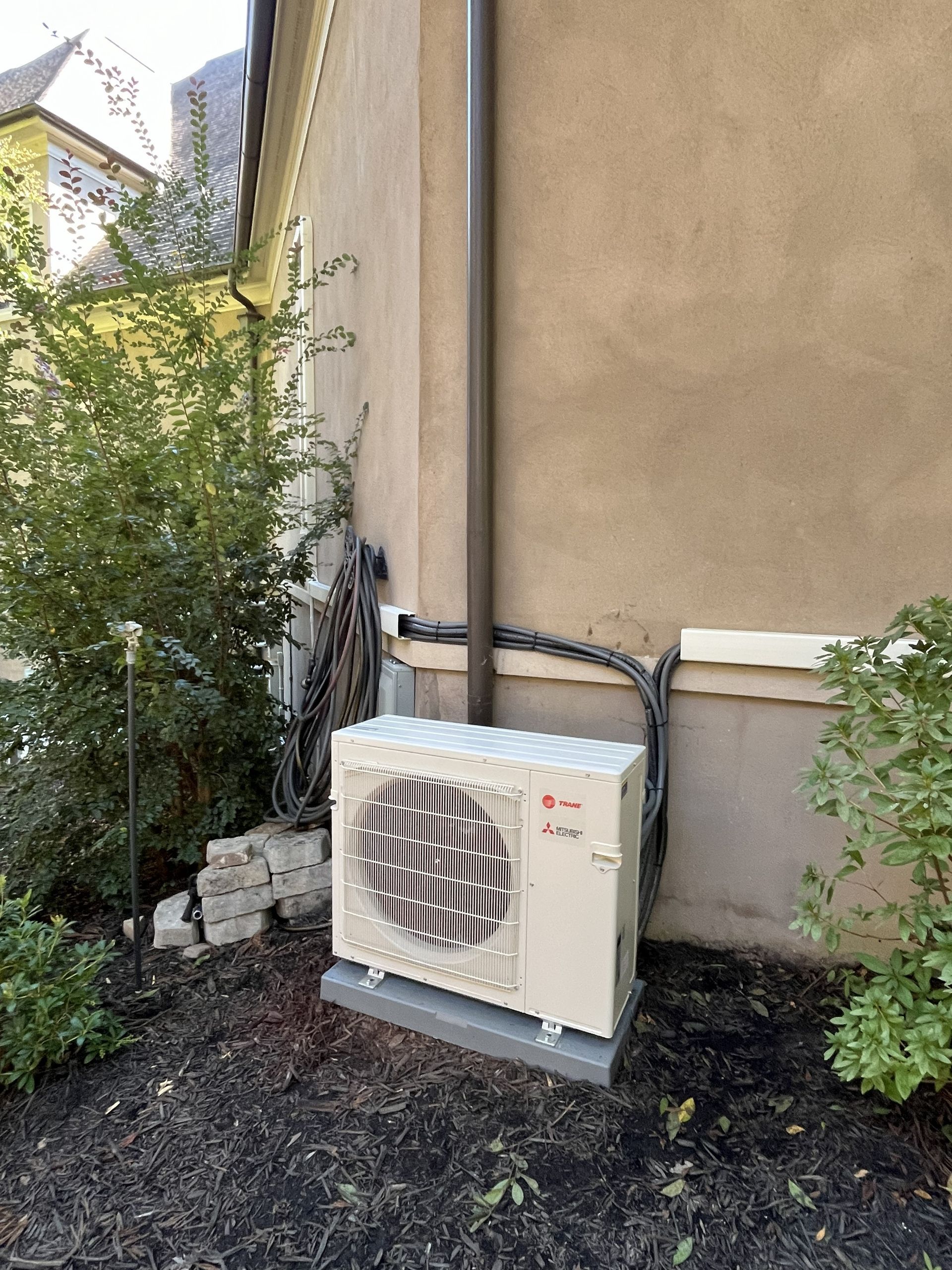 Outdoor air conditioner unit beside a beige house wall and shrubs