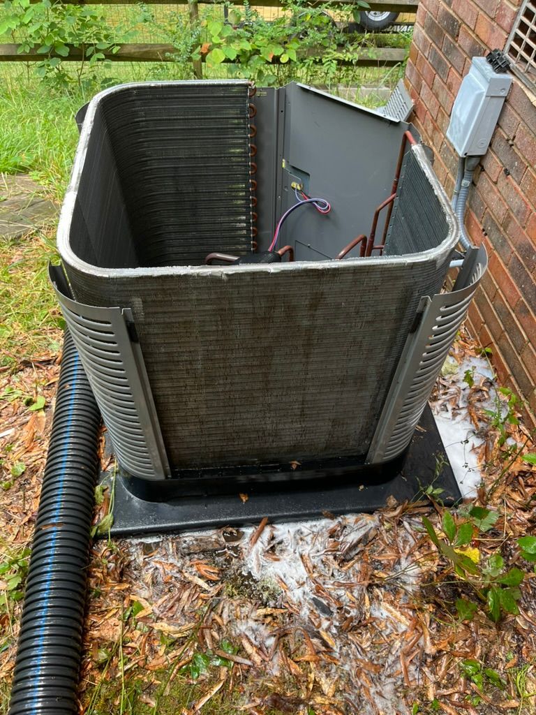 Open HVAC unit with exposed wiring beside a brick house, surrounded by wet leaves and grass.