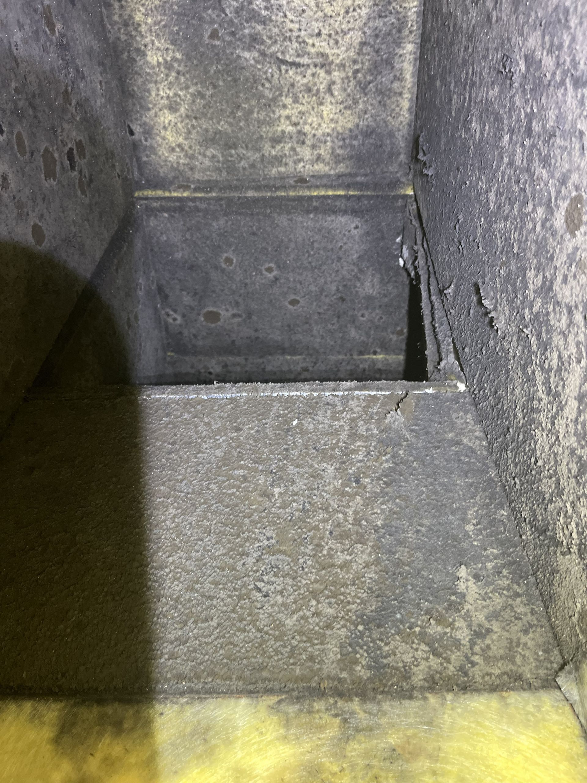 Dirty gray metal interior of a narrow bin or duct with grime and yellowed residue at the bottom