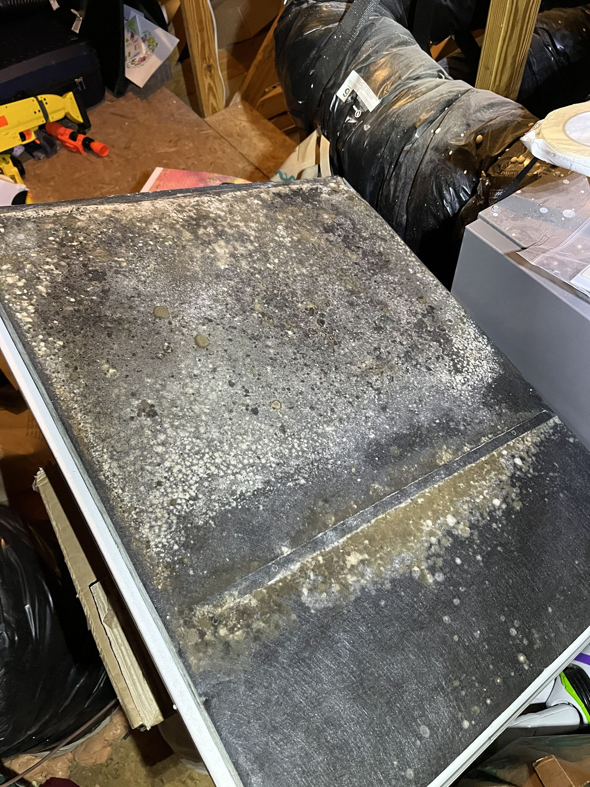 Dusty black metal vent or duct panel in a cluttered workshop, with a yellow tool and coiled hose nearby