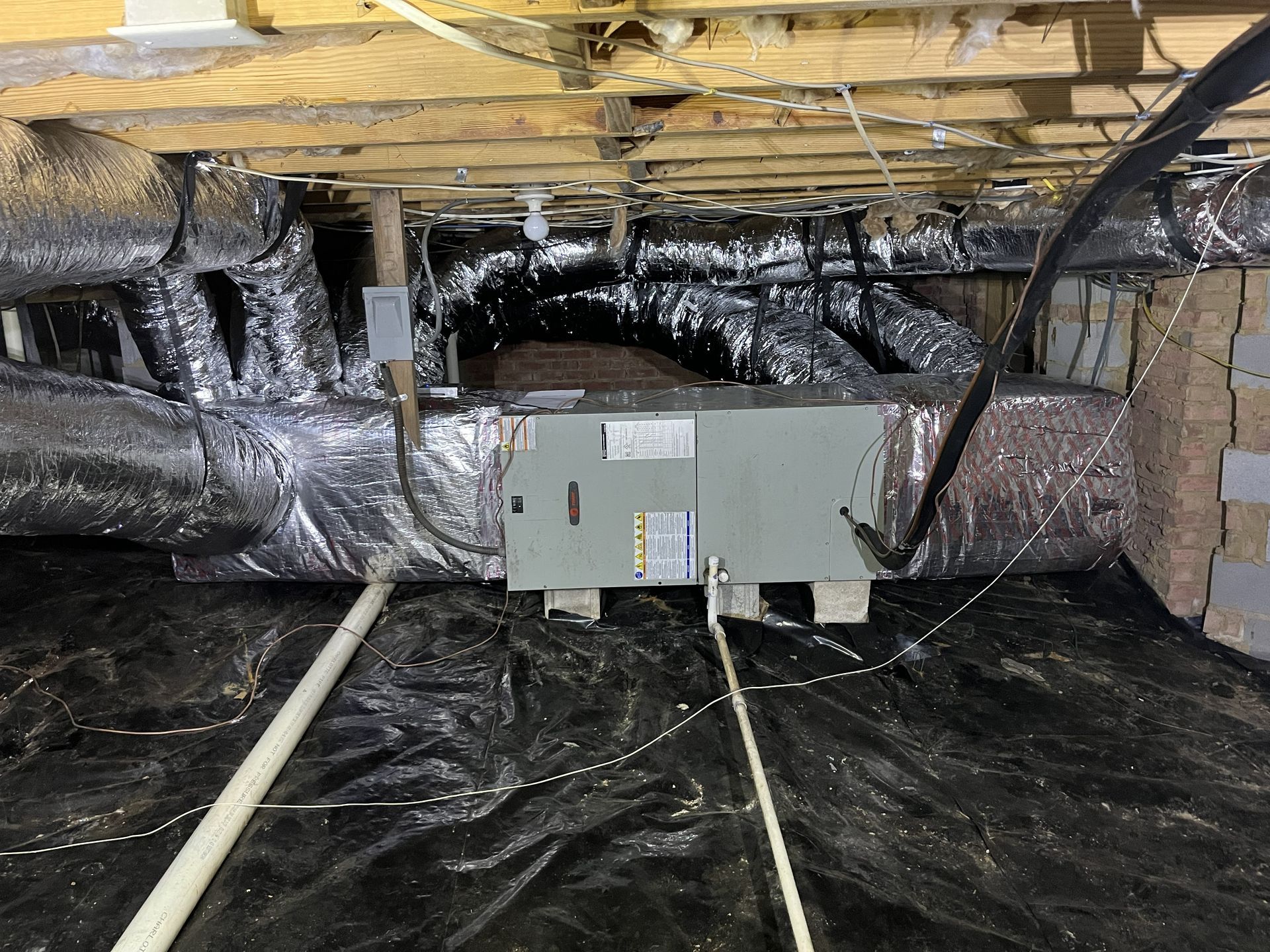 Insulated attic with a gray HVAC unit on the floor amid black plastic and exposed ductwork.