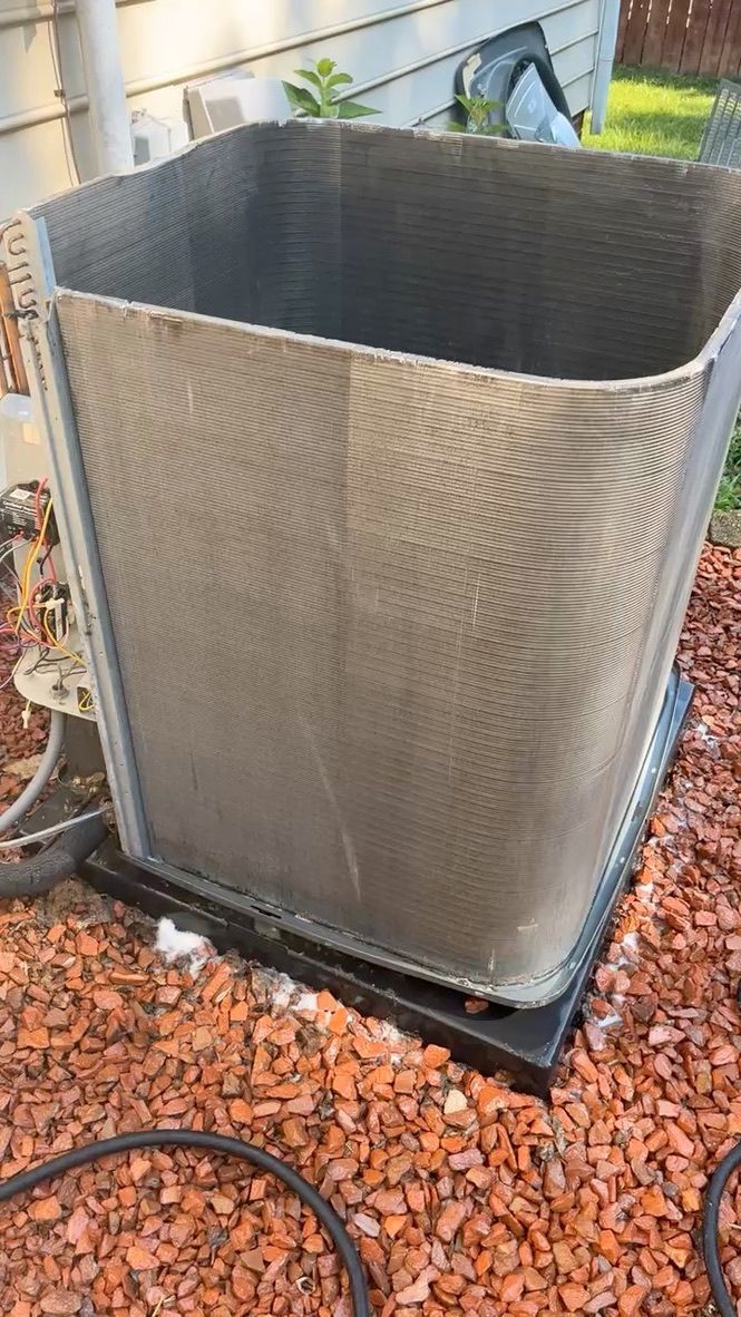 Outdoor HVAC condenser unit with silver metal grille on a black base, beside a house and red gravel mulch.