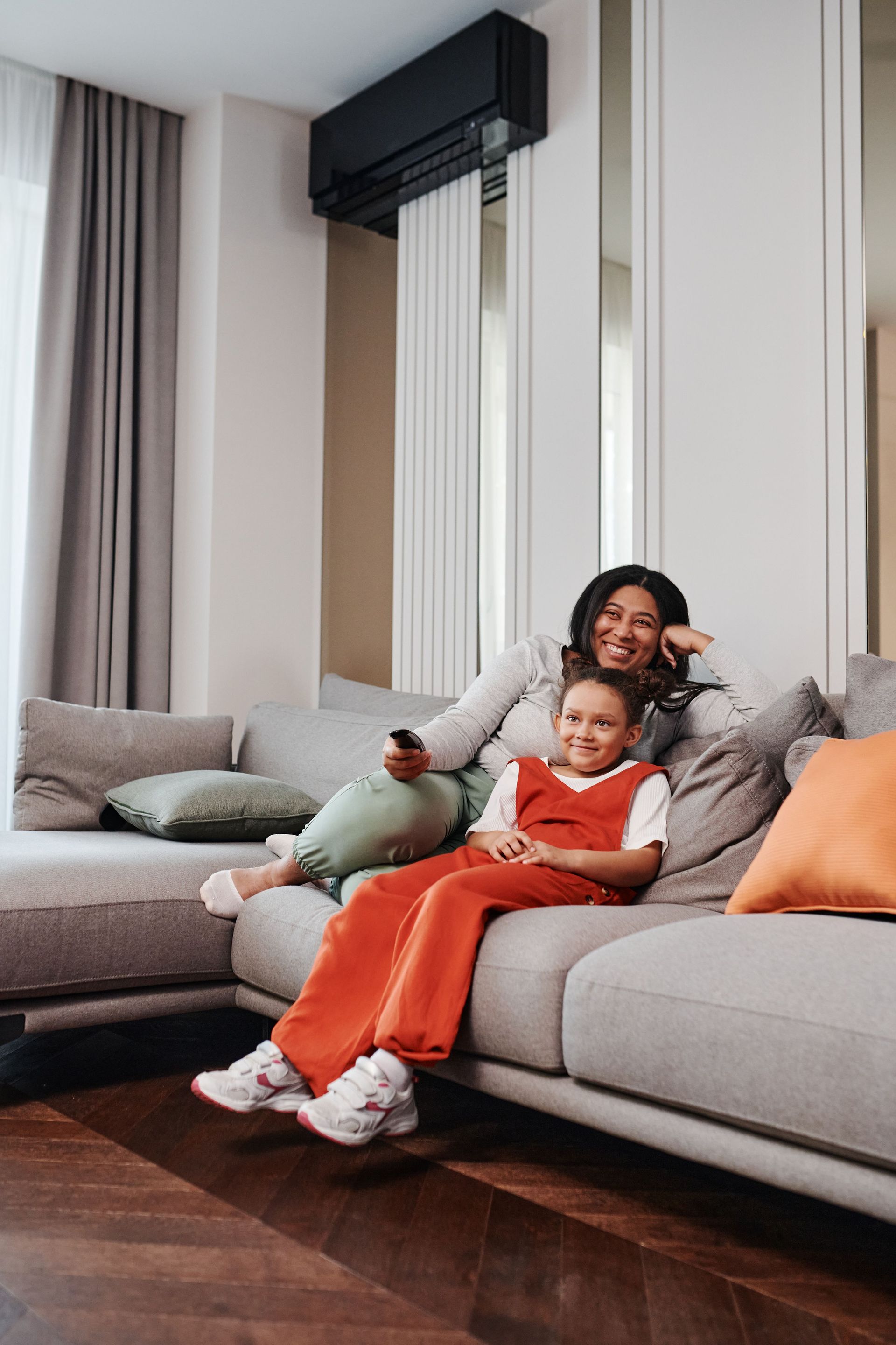 Adult and child lounging on a gray sofa in a bright living room, smiling with a remote control.