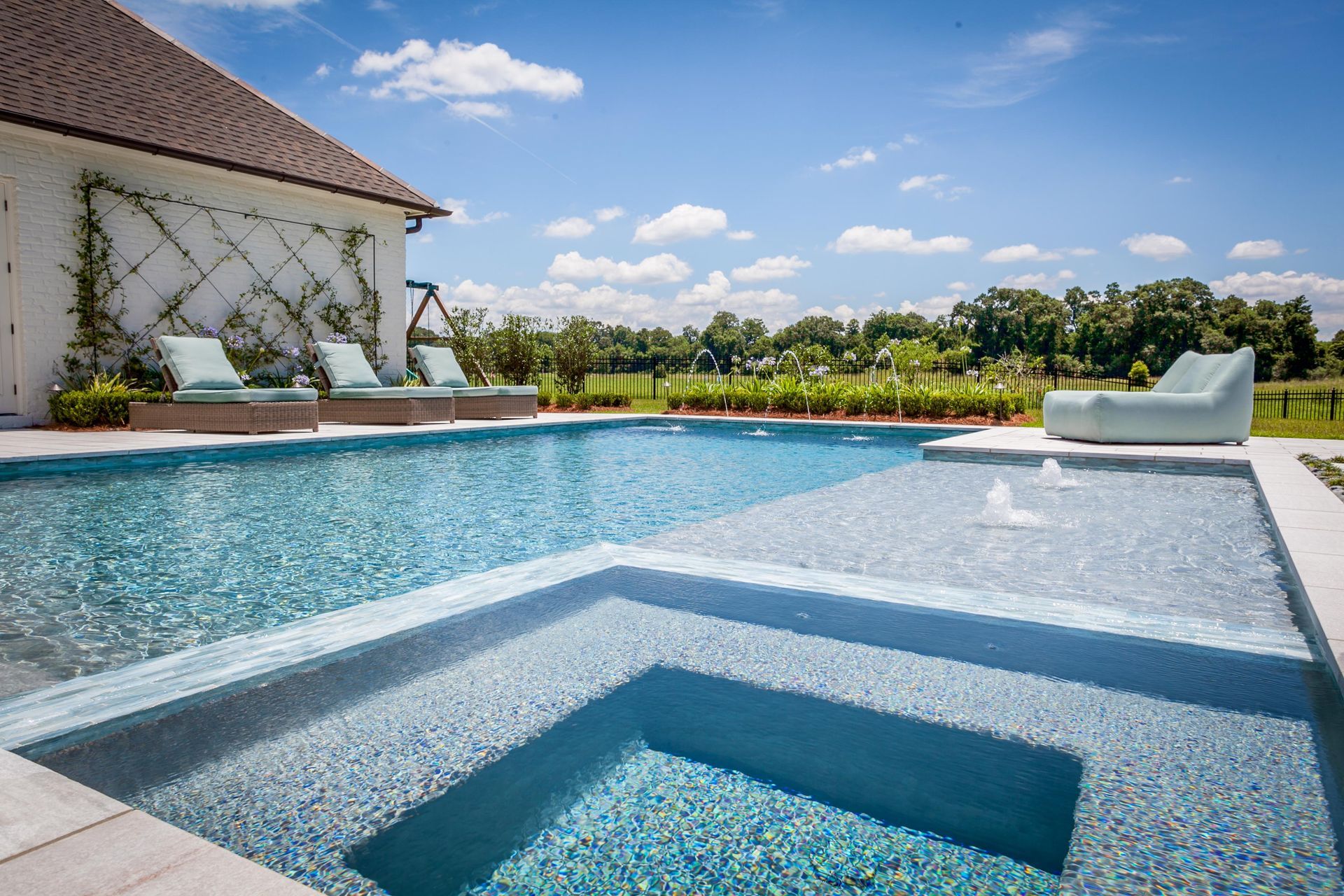 Luxurious swimming pool with a sunbathing area and a jacuzzi, next to a white building, under a bright blue sky.