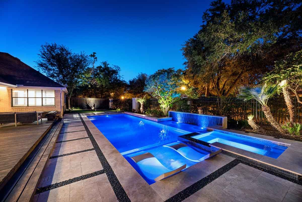 Nighttime backyard with illuminated blue swimming pool, spa, and surrounding patio. Trees and a house are visible in the background.