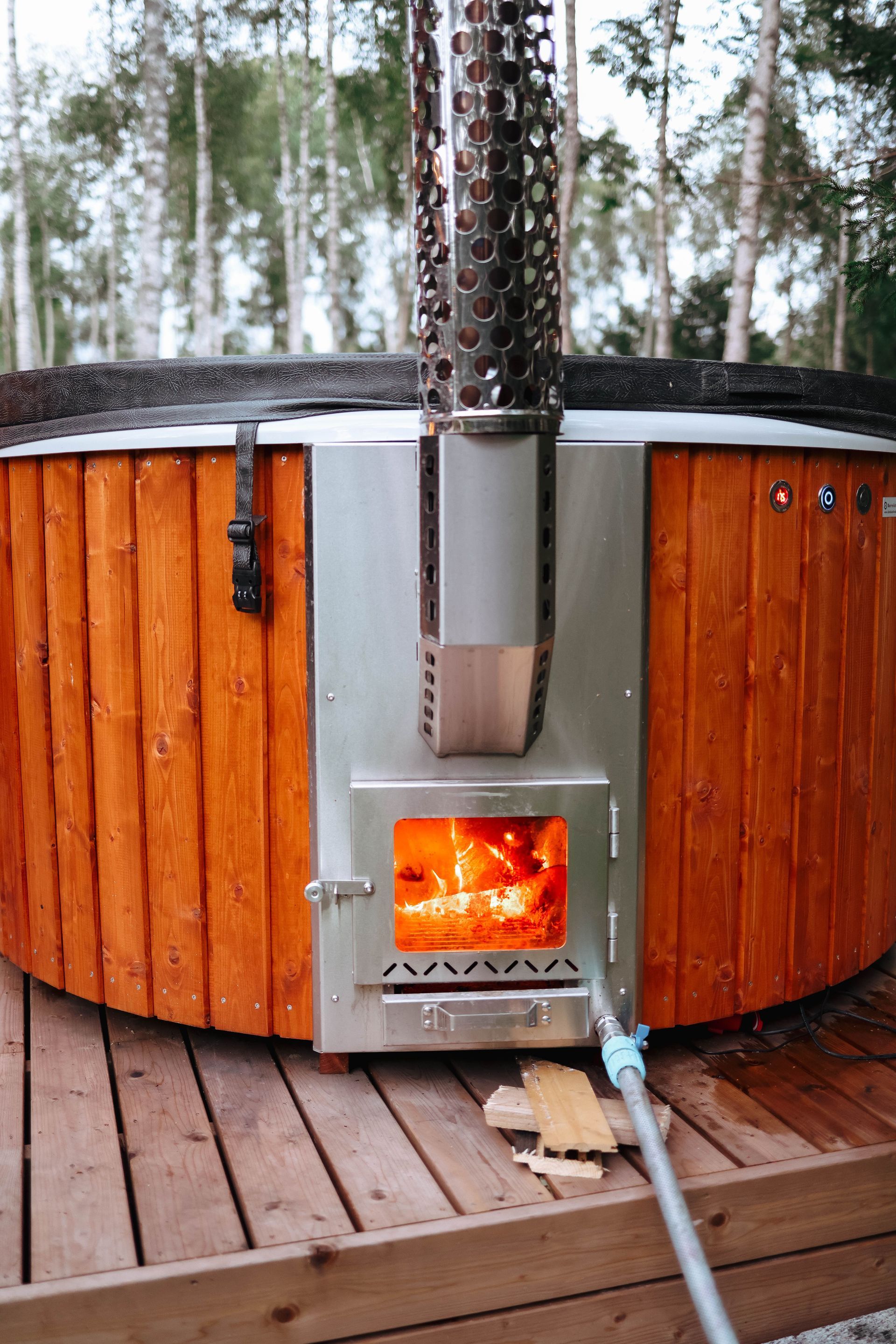 Wood-fired hot tub with a lit stove and metal chimney on a wooden deck outdoors