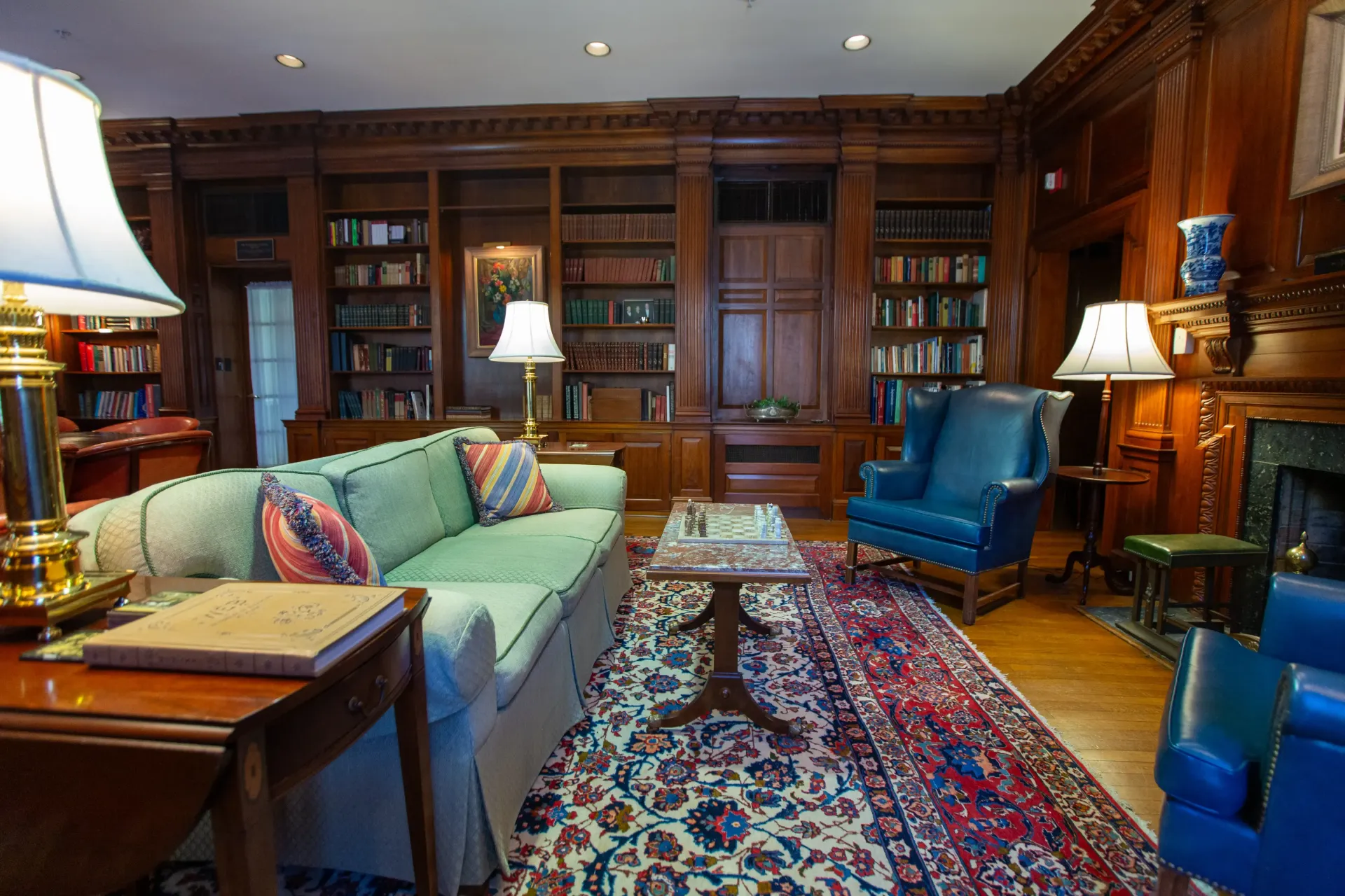 A wood-paneled library with a green sofa, blue armchairs, and bookshelves filled with books.