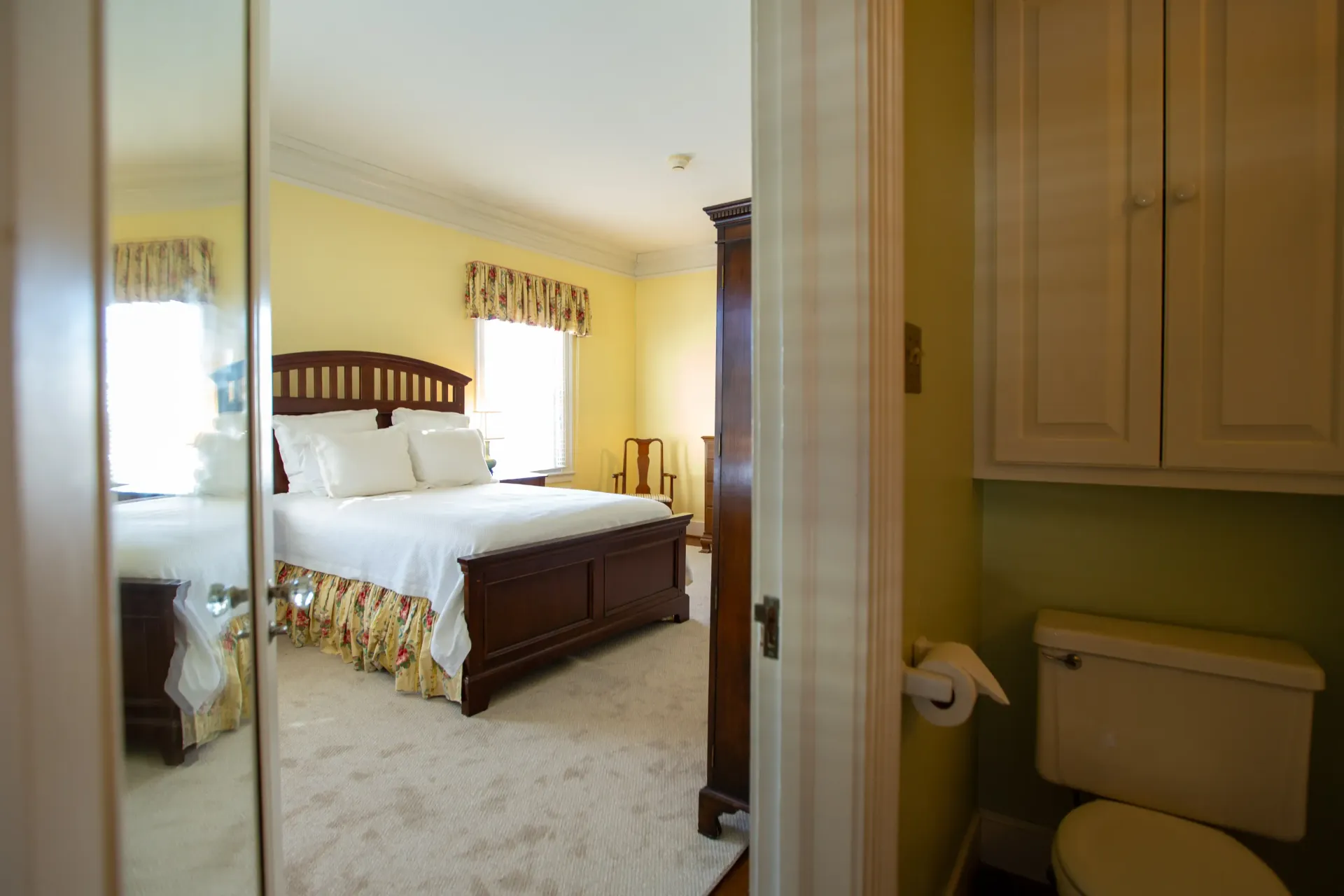 View through doorway of a bedroom with a bed and a bathroom with a toilet and cabinet.