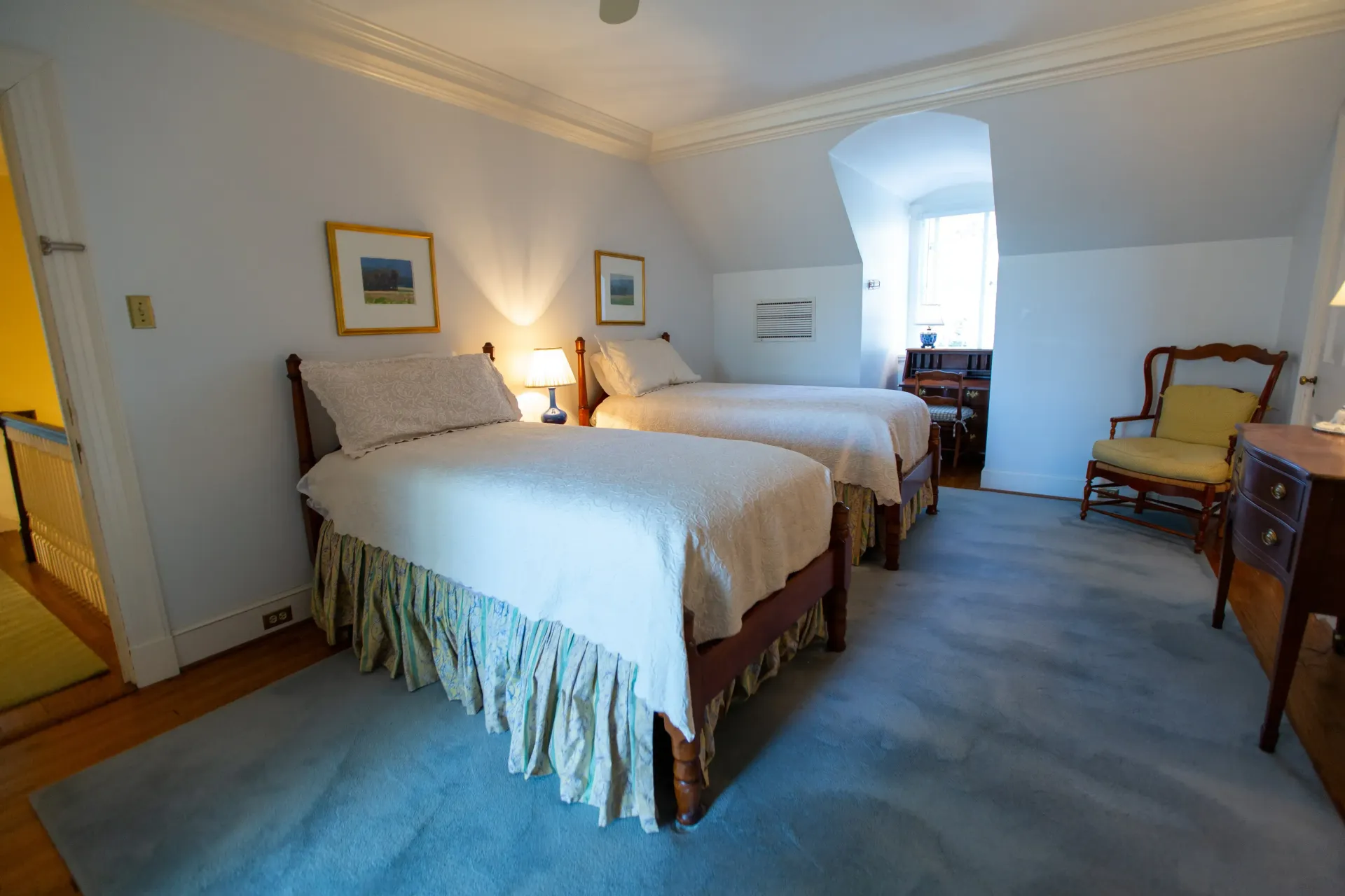 Two twin beds in a bedroom with a blue rug, two framed pictures, and a side table.