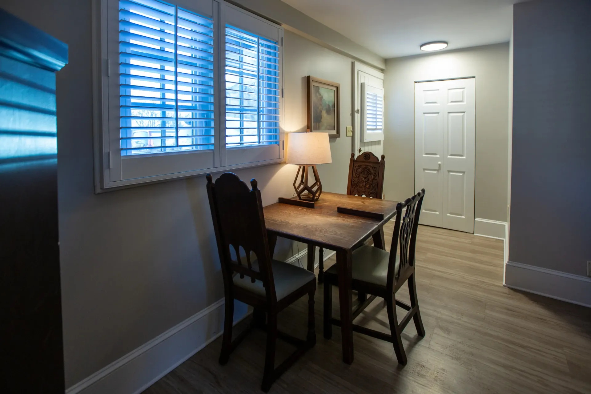 Small dining table and chairs near window with shutters, in a room with a door.