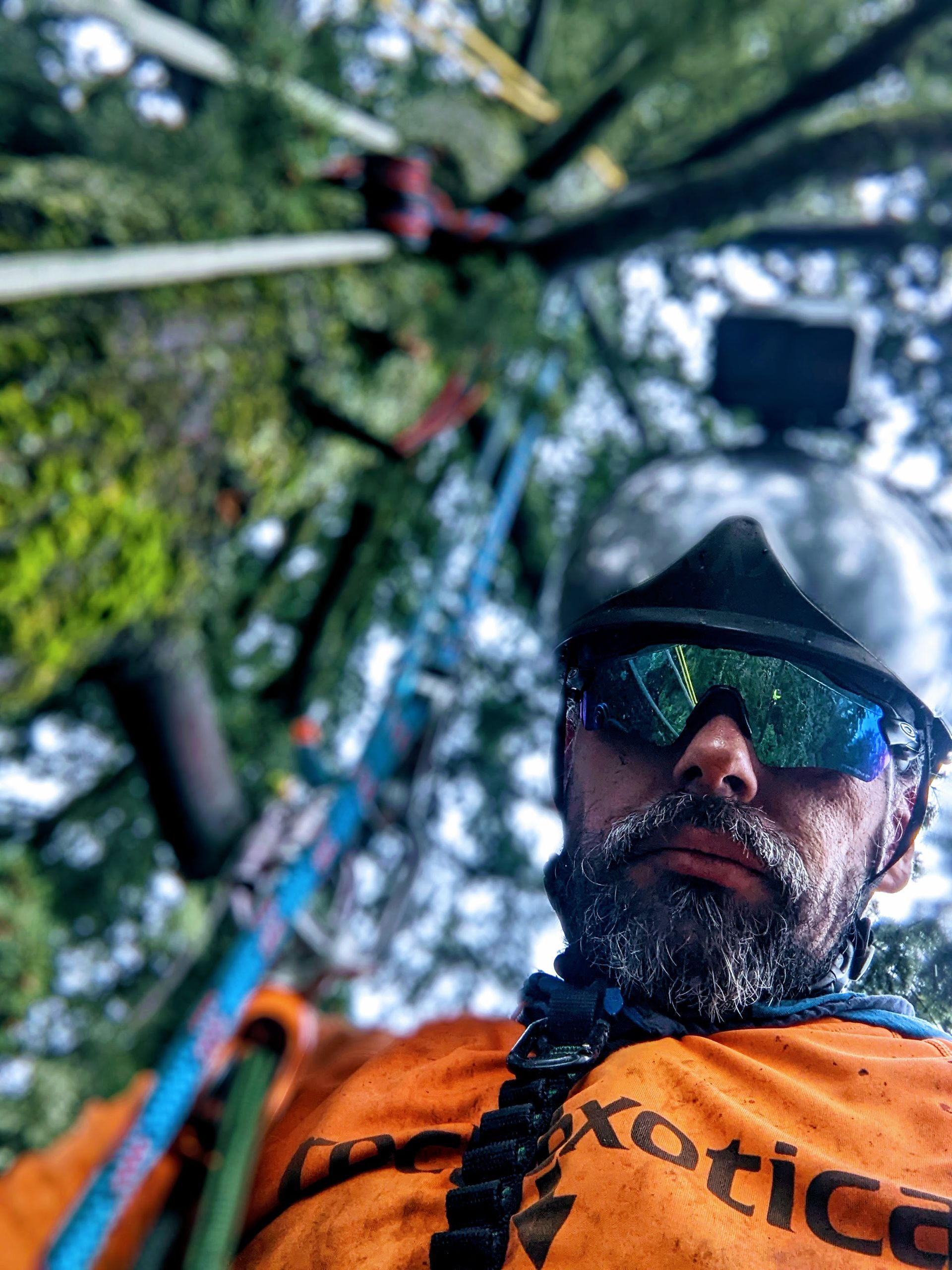 Man in orange jacket and sunglasses, seen from below in a forest canopy with a small aerial camera above.