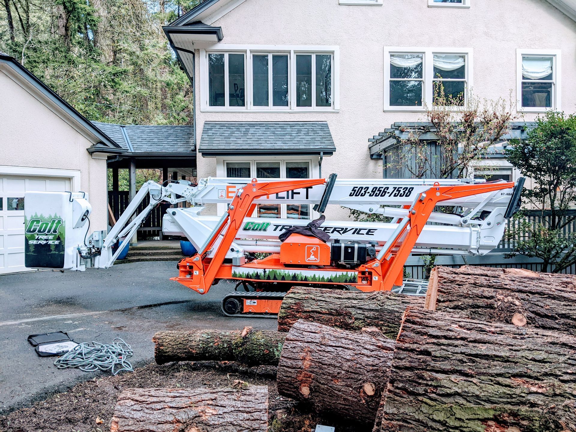 Orange telescopic boom lift at a house under construction, with logs and tree debris in the foreground.