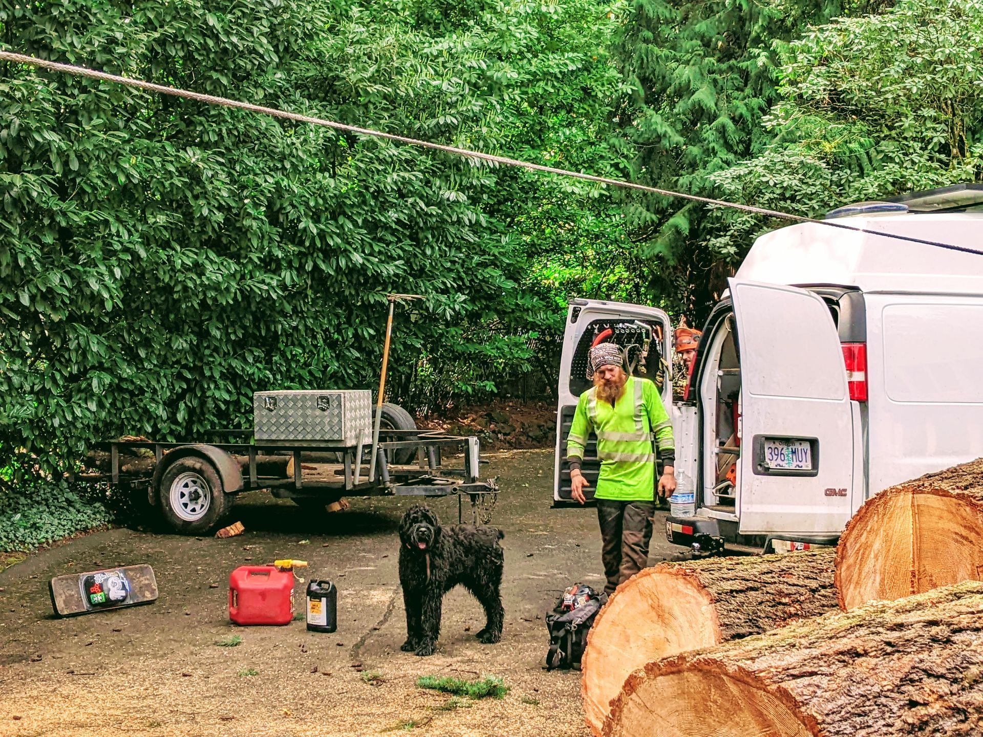Worker in neon jacket beside a white trailer and black dog in a muddy wooded campsite with tools.