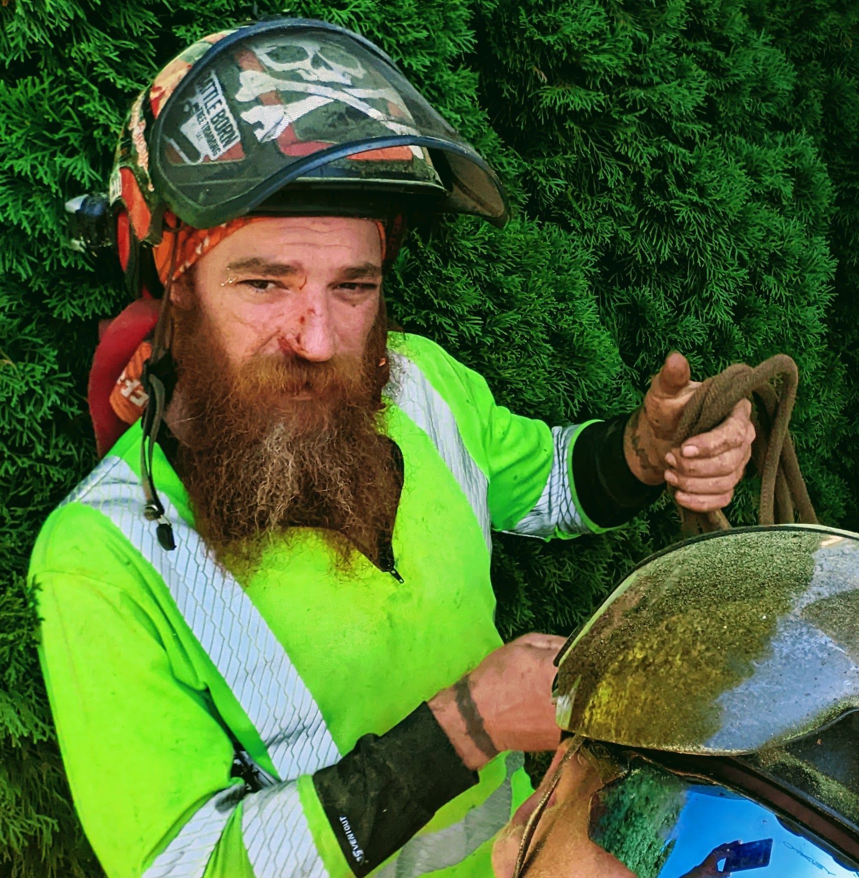Man in a neon safety jacket and helmet holding a motorcycle helmet beside green hedges