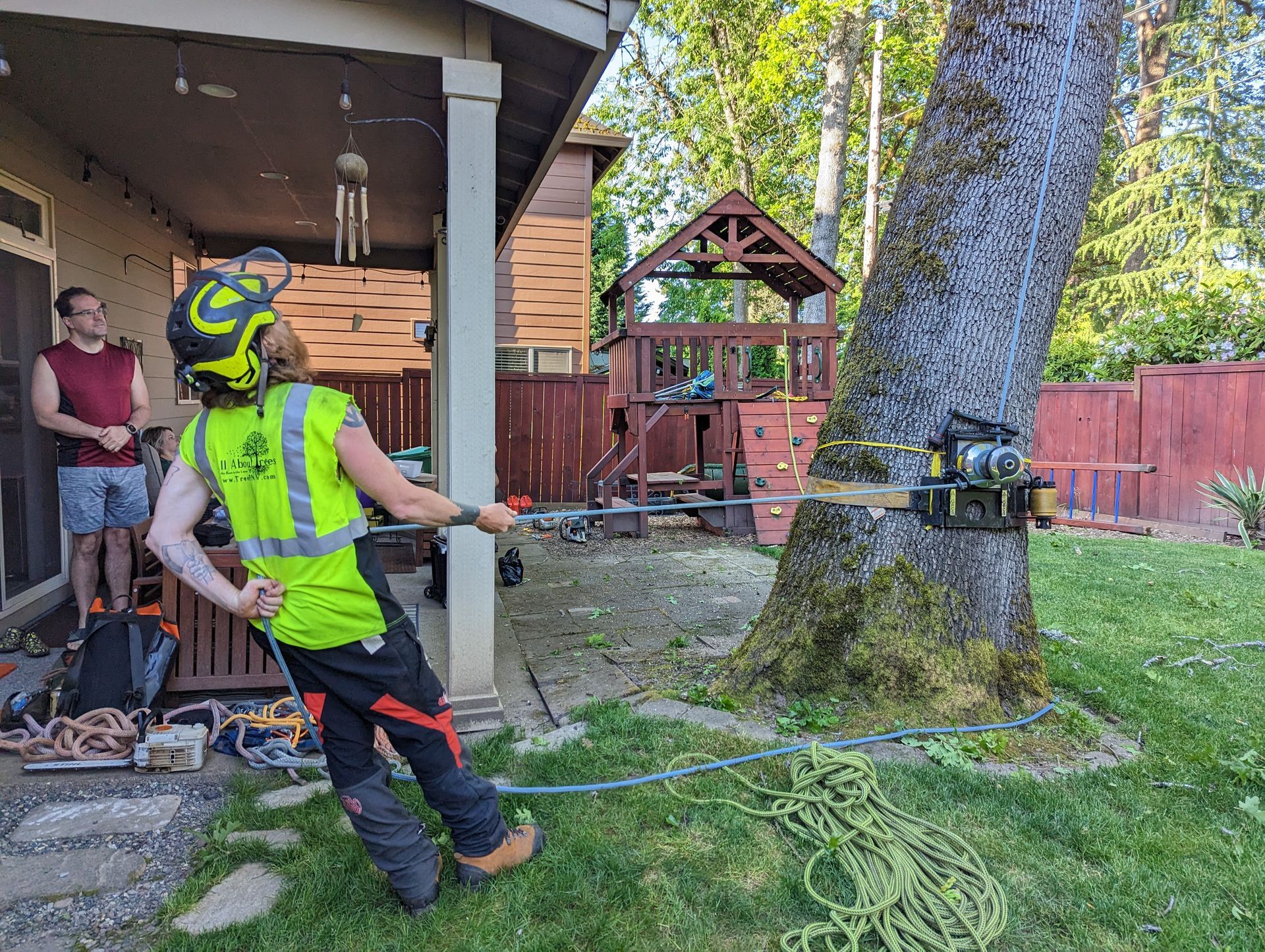 Person in neon safety gear uses a rope around a large tree in a backyard near a house and playset.