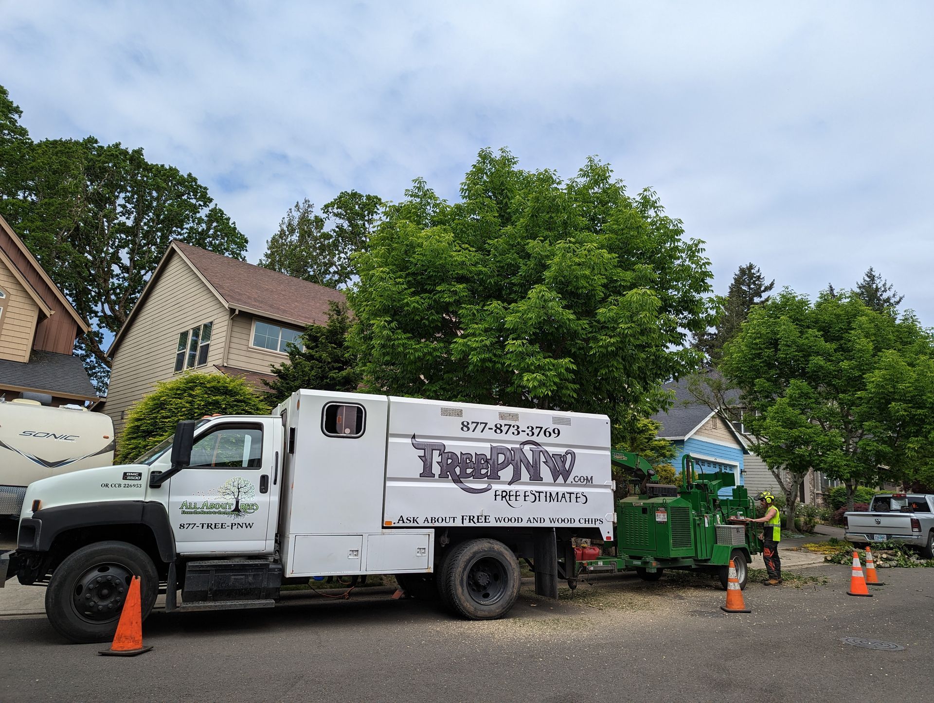 Work truck and green tree-service machine parked on a residential street, with orange cones nearby