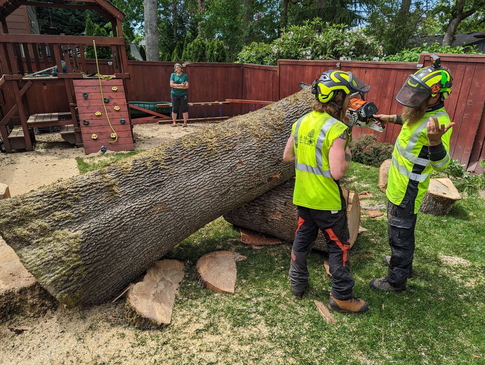 Workers in neon safety vests inspect a felled tree in a backyard, with one person standing nearby.
