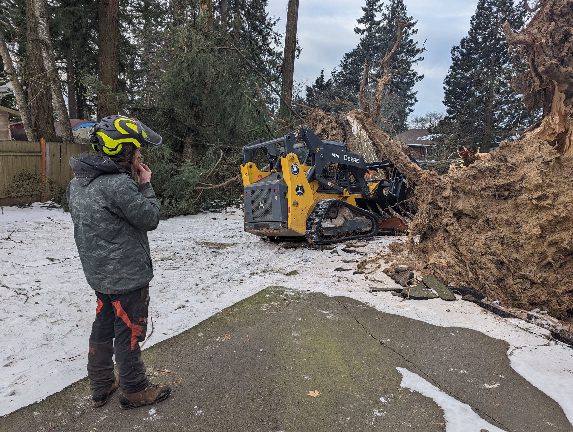 Worker in neon helmet filming a yellow skid steer clearing a fallen tree on a snowy driveway