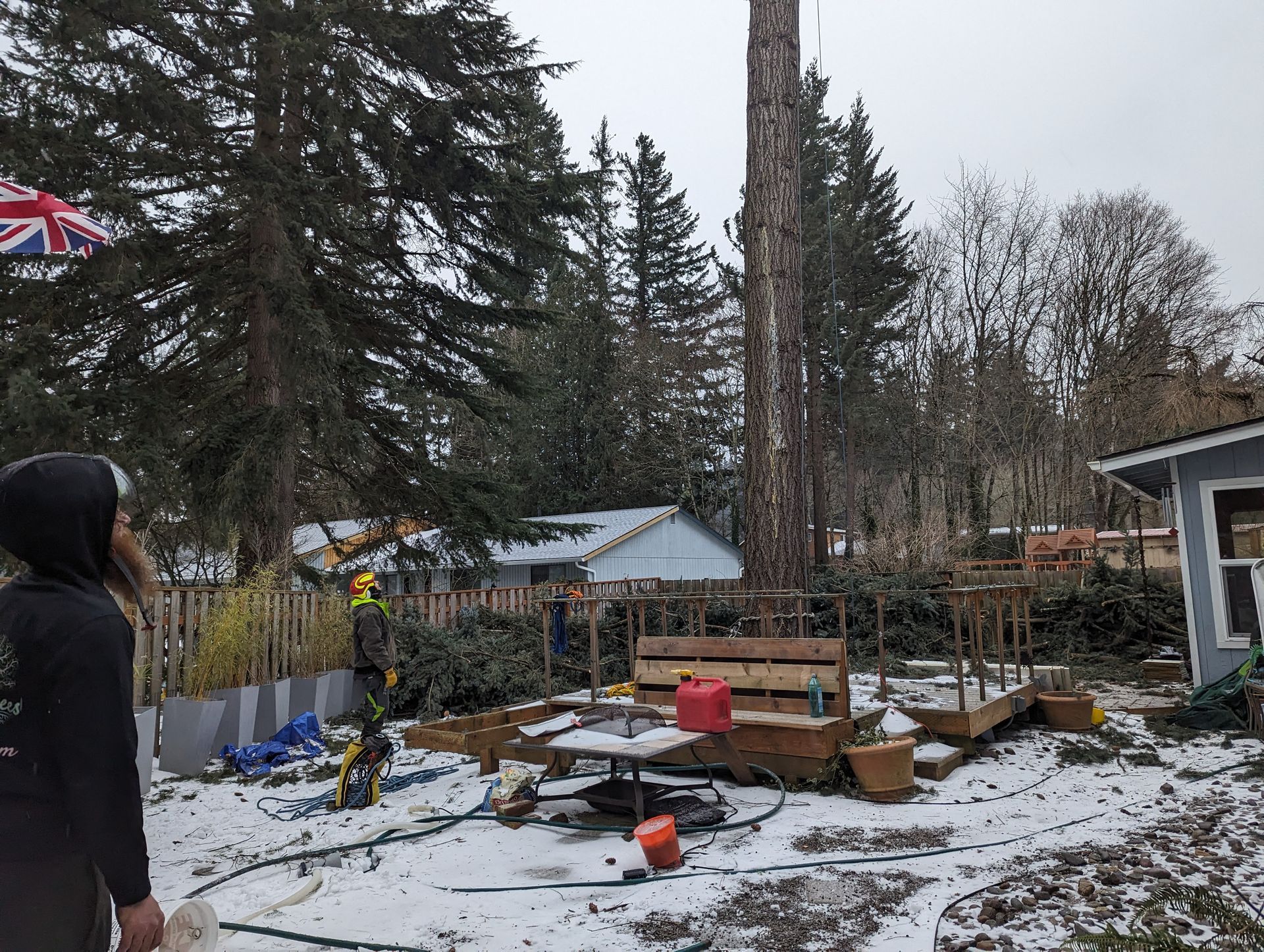 Snowy backyard with scattered debris, a person standing at left, and tall trees and houses in the background