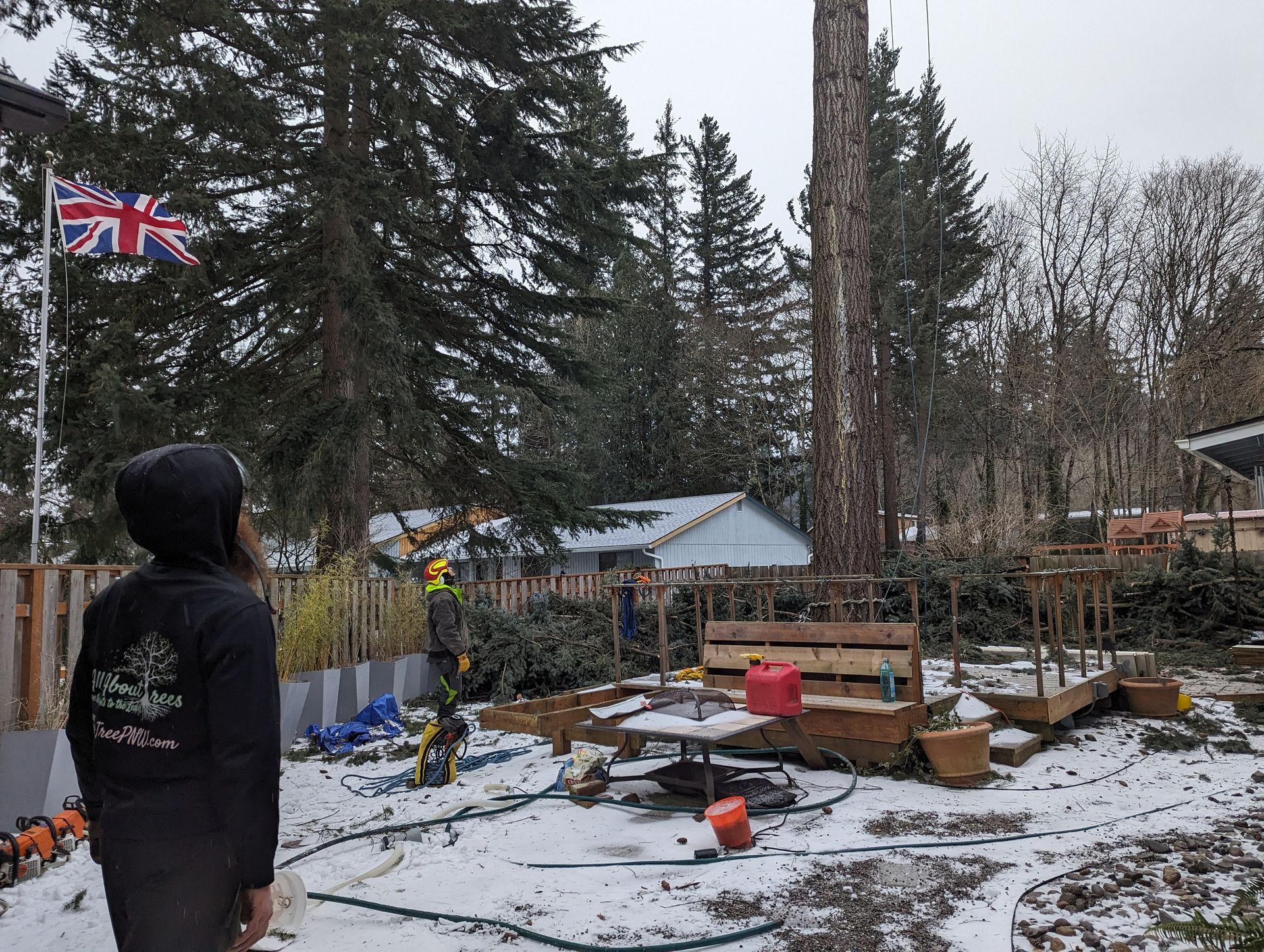 Person in a black hoodie stands in a snowy yard with garden beds, trees, and a small flag.