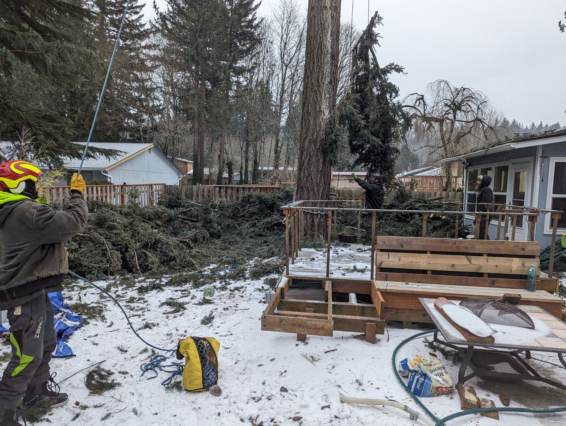 Snowy backyard with a person near a fire pit, benches, and a wheelbarrow by a fence and house
