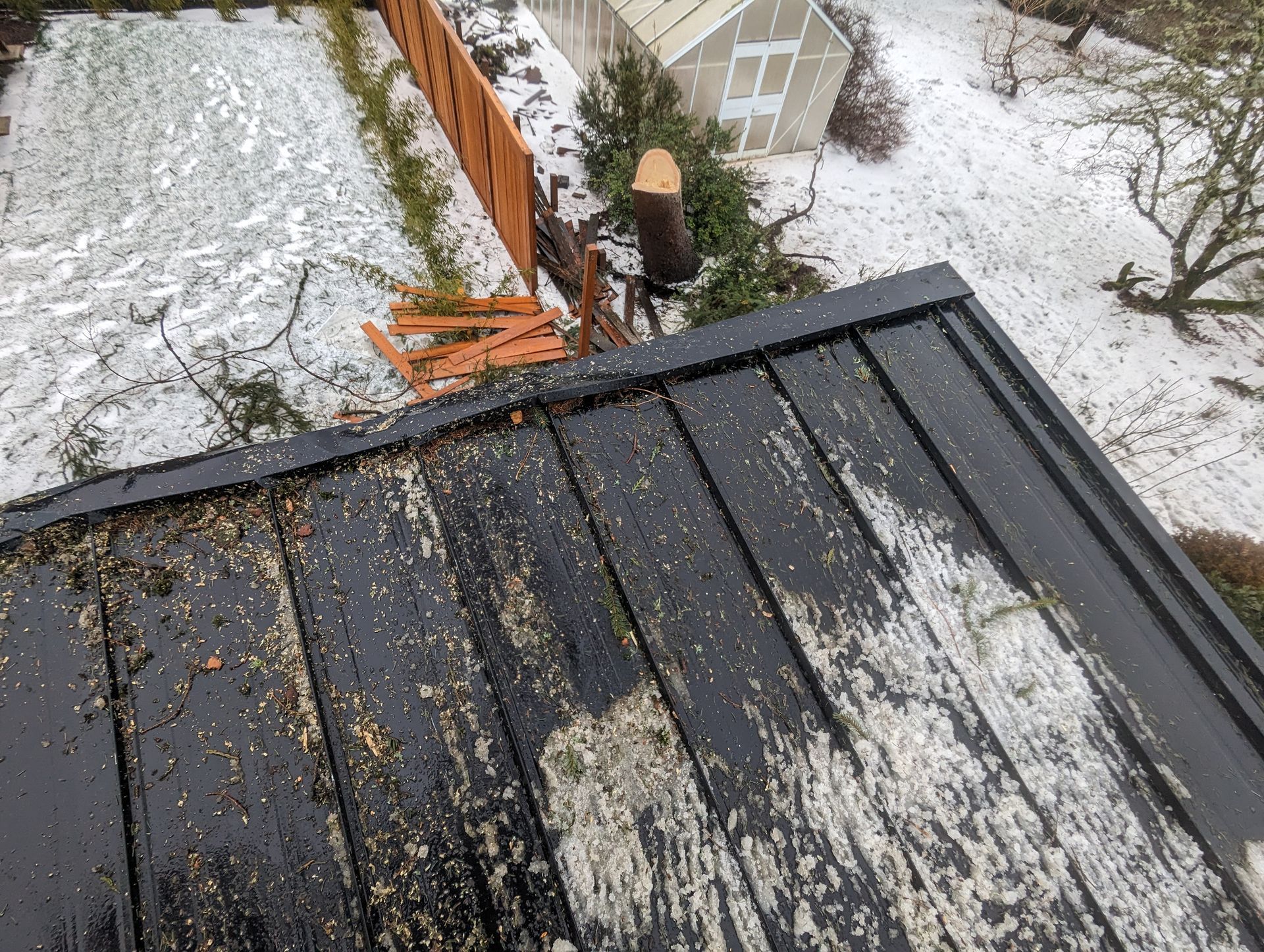 Snow-covered rooftop with a person clearing debris near a fence and a small shed below