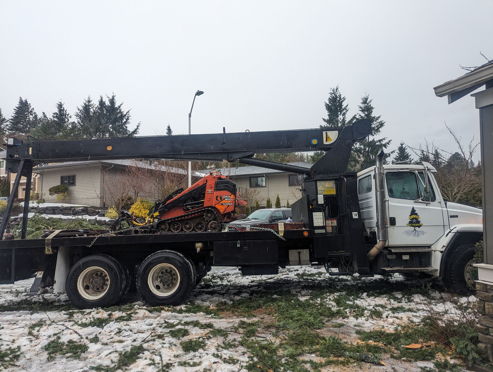 White flatbed tow truck carrying an orange skid steer in a snowy yard under overcast skies