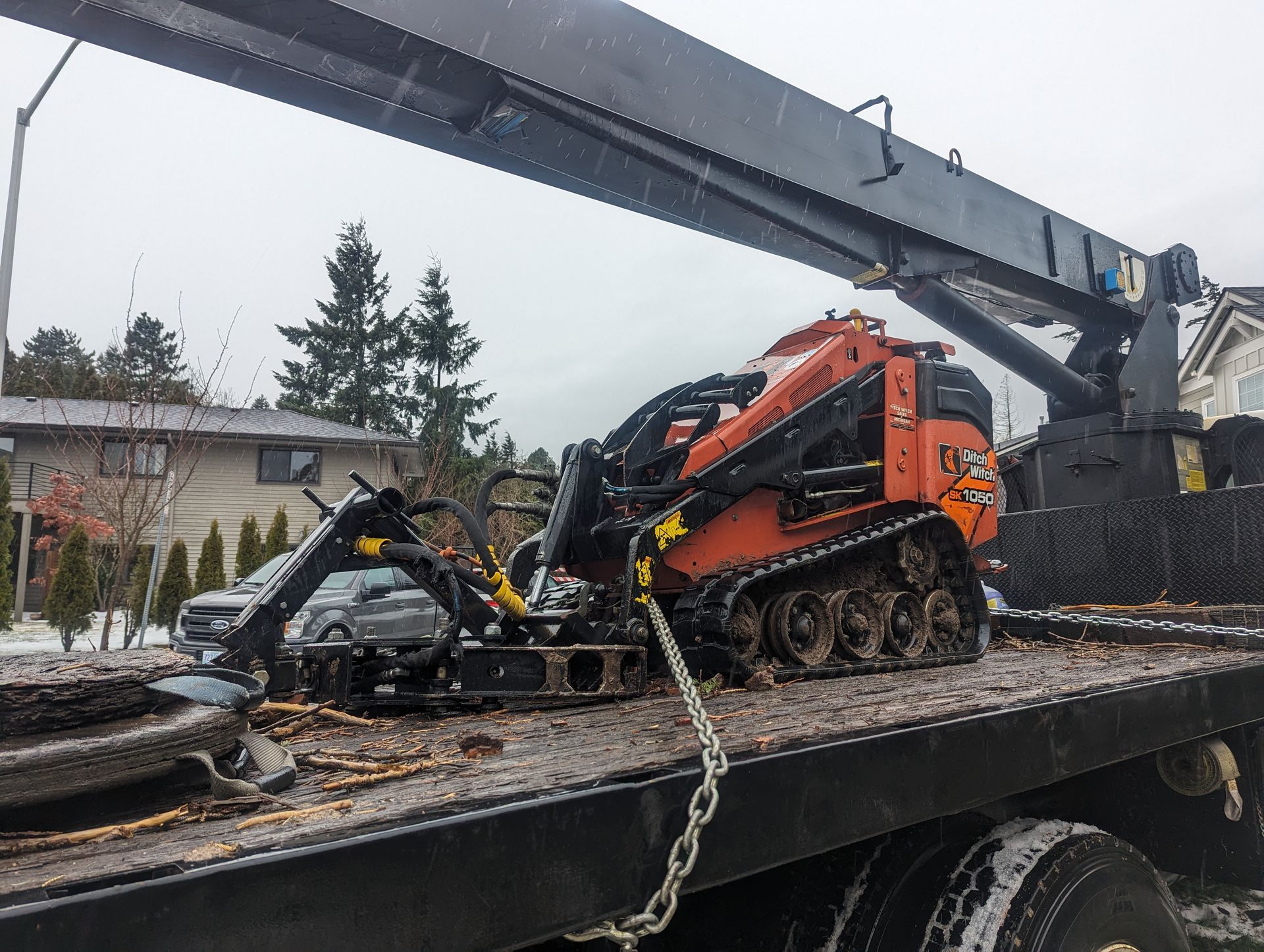 Orange tracked construction machine on a flatbed truck beneath a large crane arm in a snowy worksite