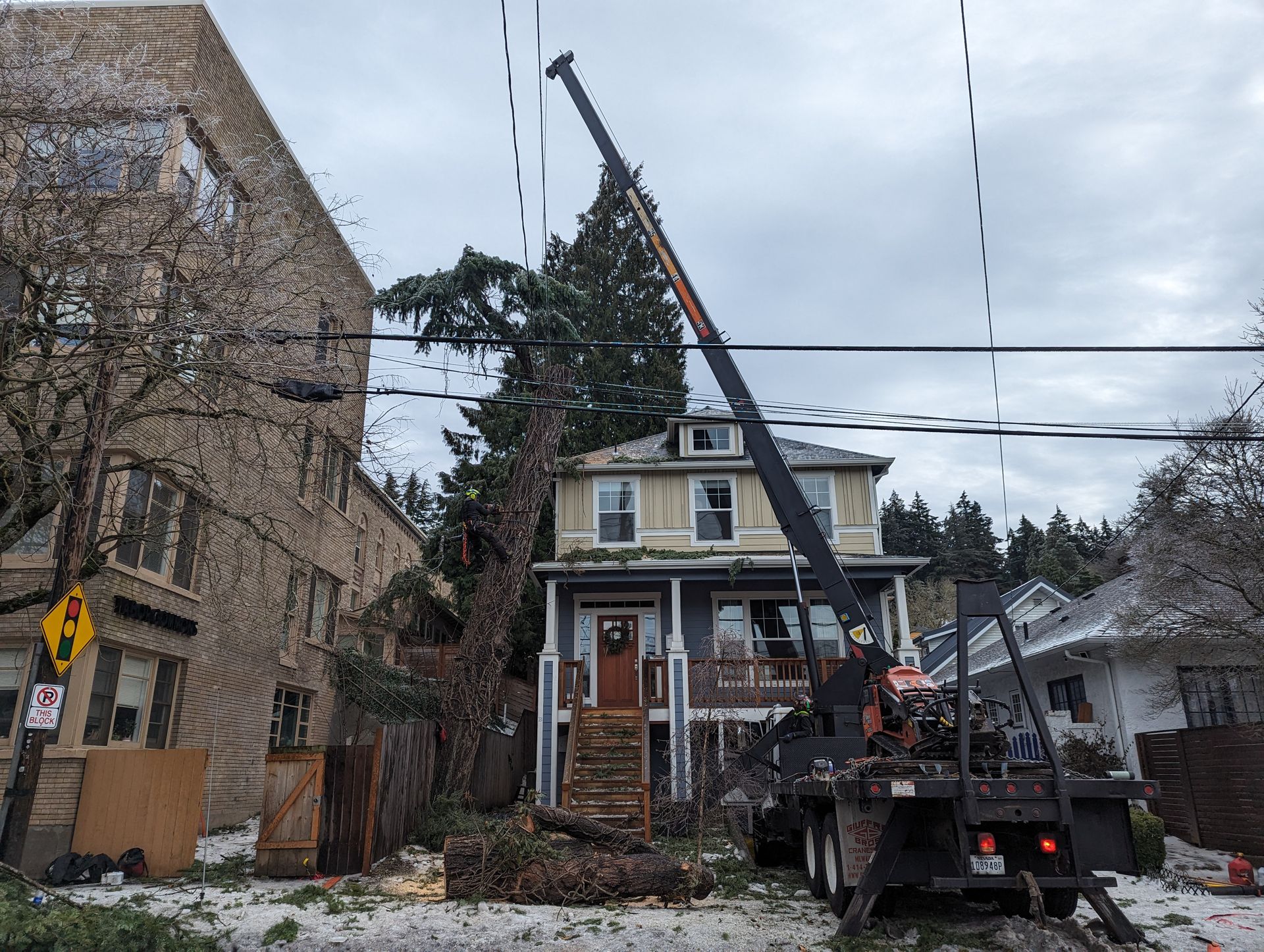 Workers near a house with a steep roof, using a truck and crane on a cloudy street.