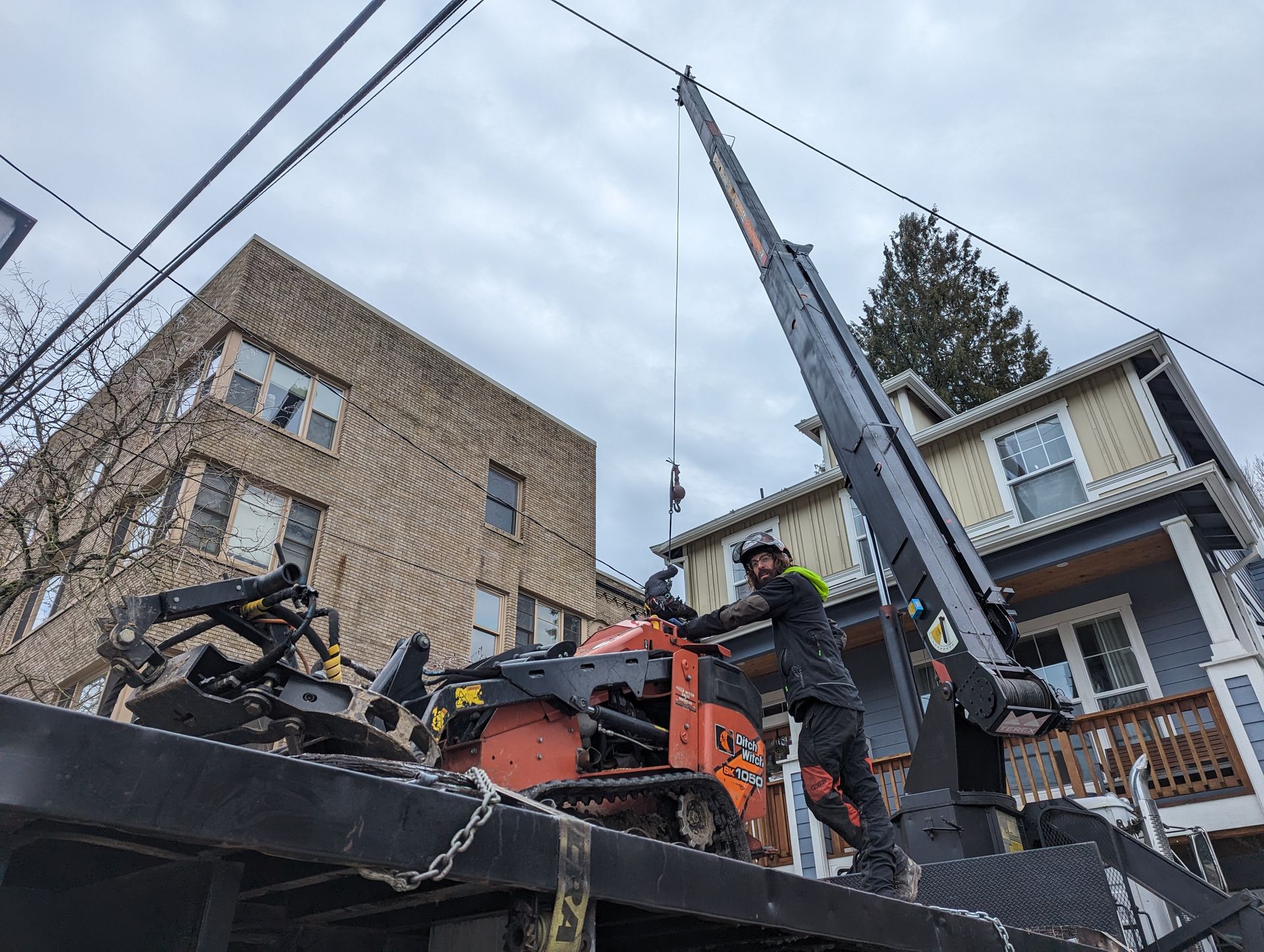 Worker operating an orange crane near damaged houses and power lines after a collapse.