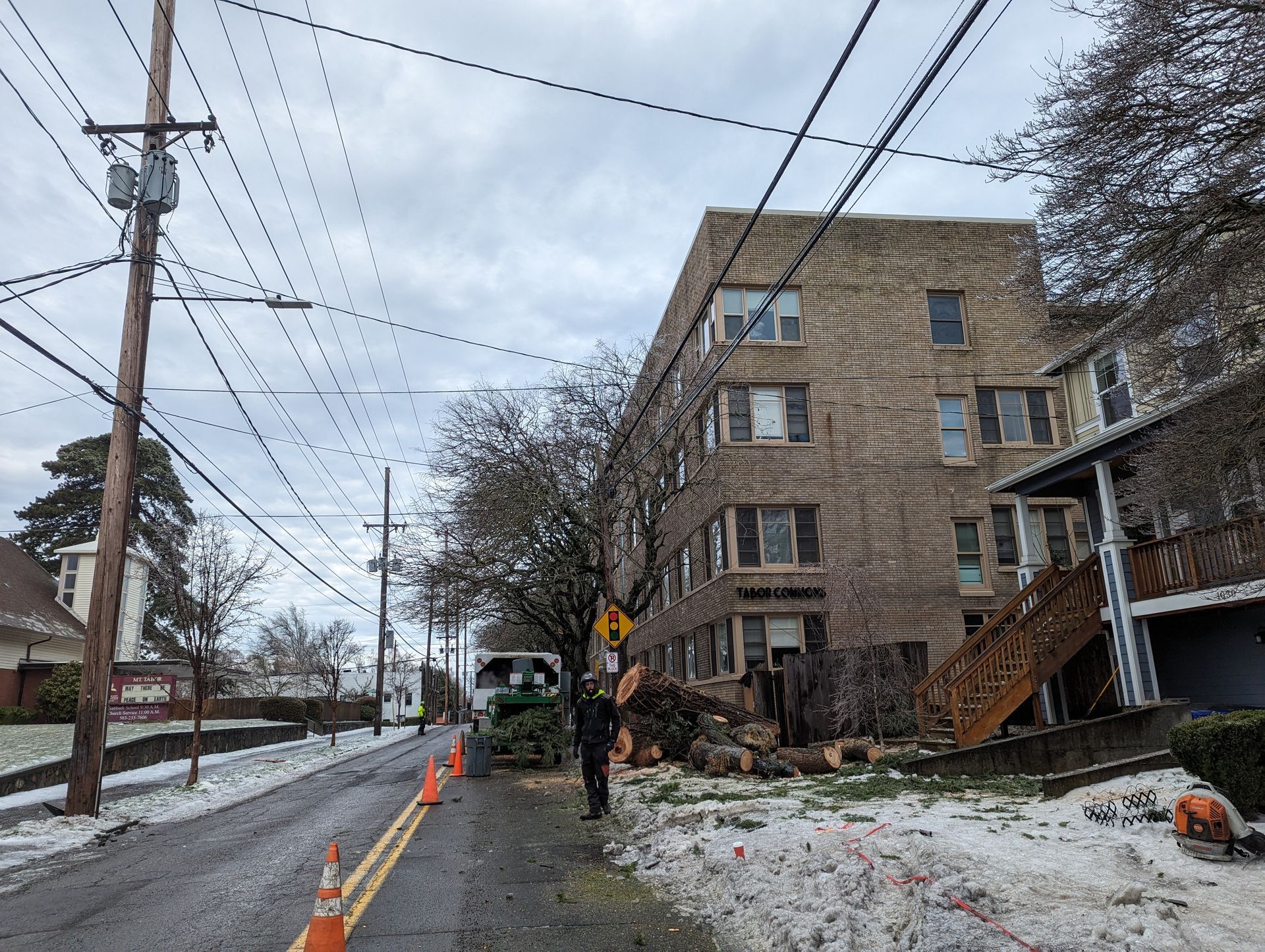 Snowy street with a damaged apartment building, utility poles, and roadside caution cones after a storm.