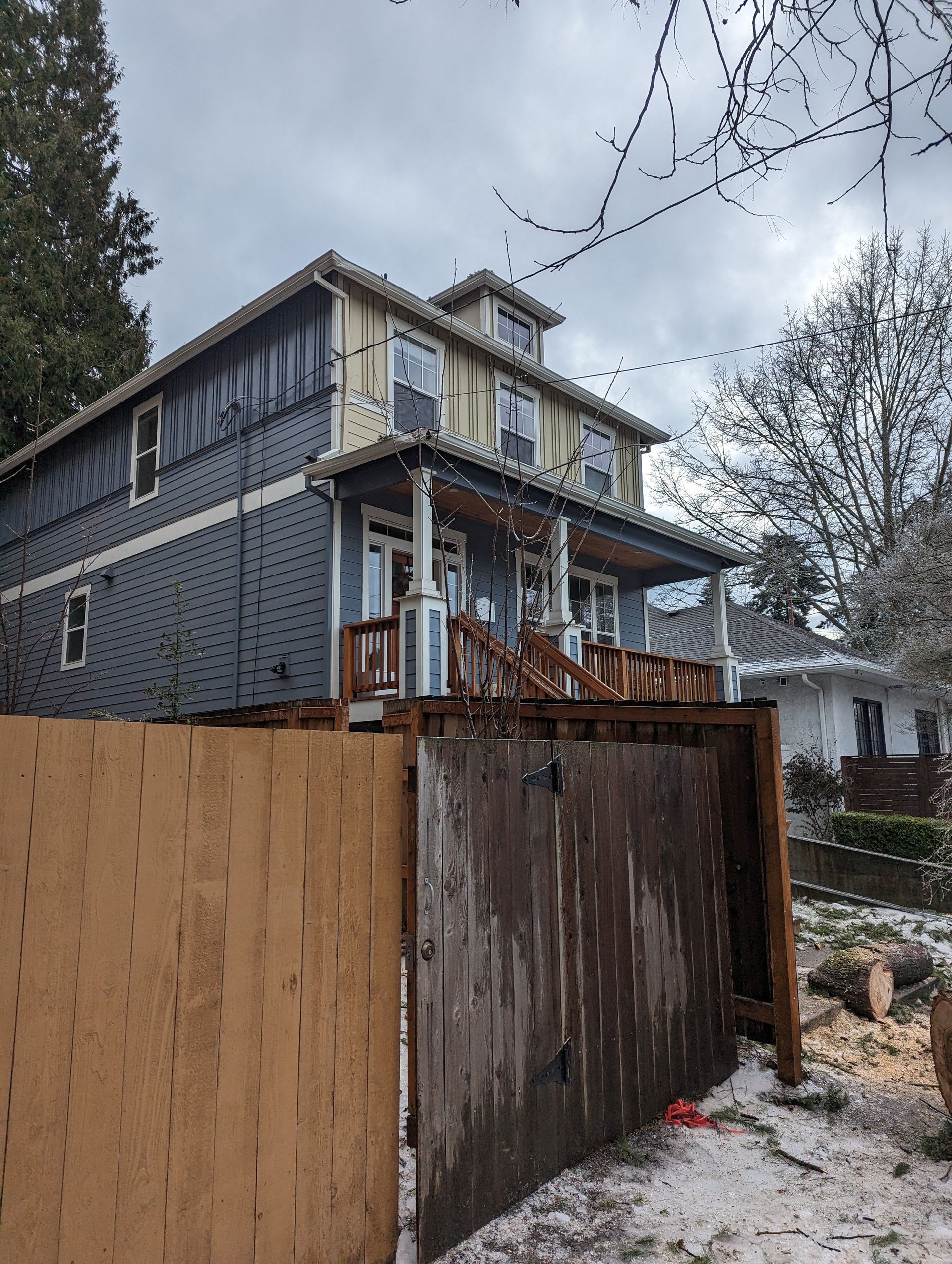 Two-story house with a wooden deck and tall privacy fence in a snowy yard under cloudy skies