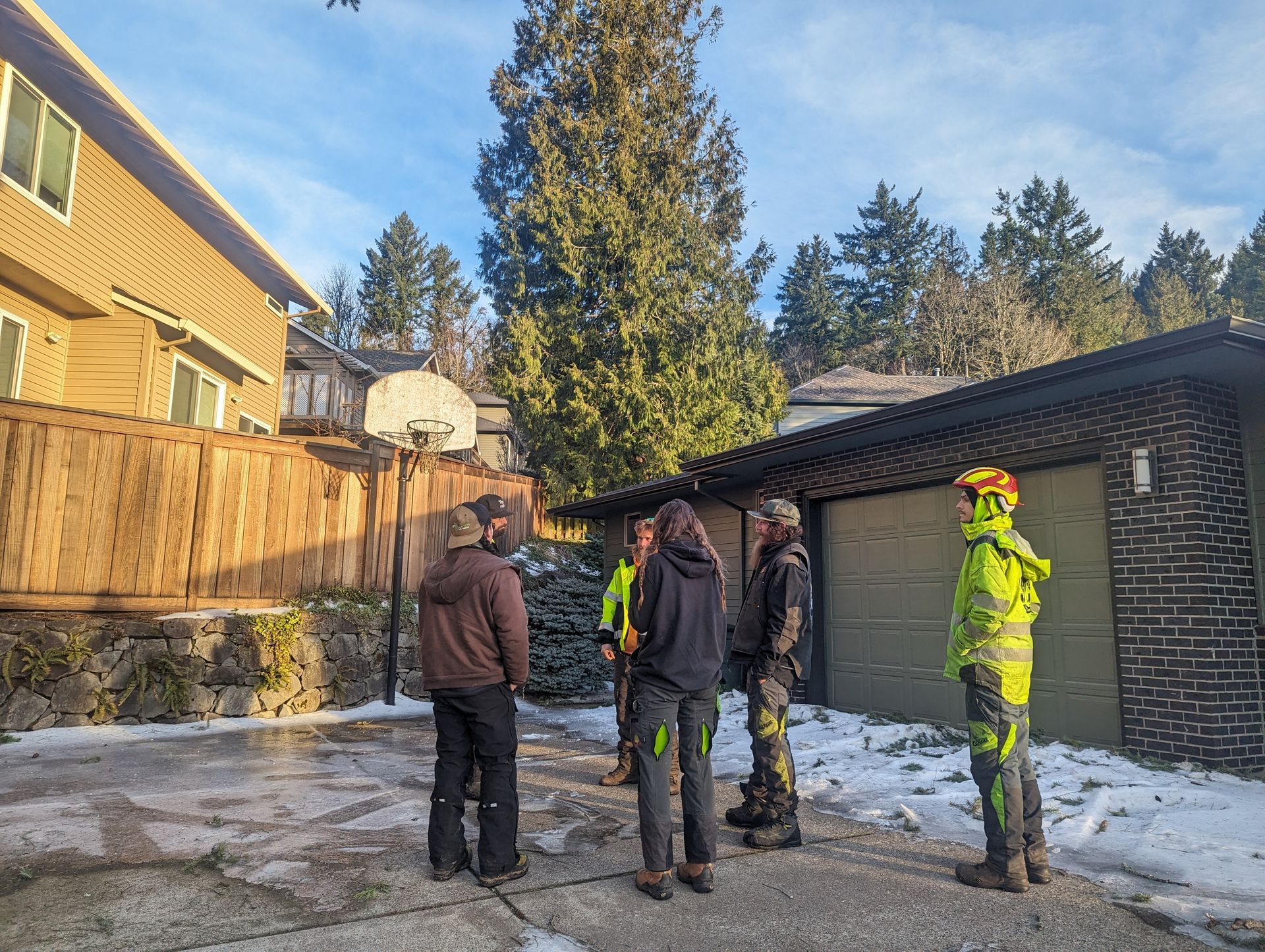 People in winter coats stand in a snowy backyard beside a garage and wooden fence.